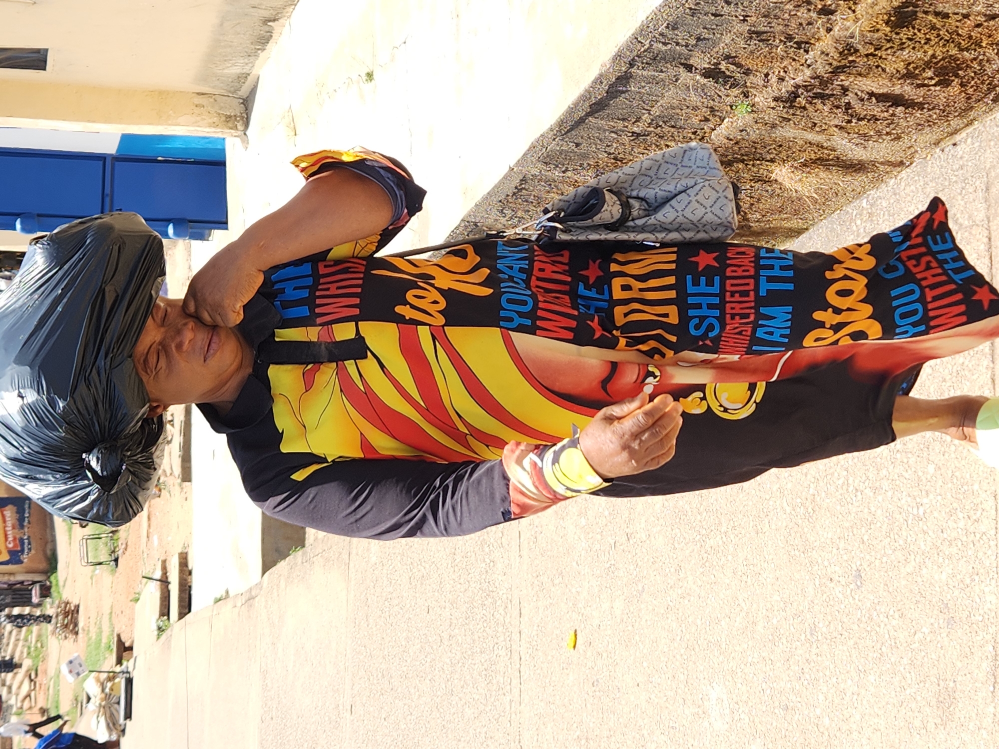 A woman in a black and patterned dress with a black plastic bag on her head stands on a concrete surface. She appears to be holding a small object in her hand. Behind her, a building with blue windows is visible. In the background, there's some vegetation and what appears to be construction or discarded materials. The words "THE WHIS," "YOU CAN'T," "SHE WHISPERED BACK," and "Star" are printed on her dress.