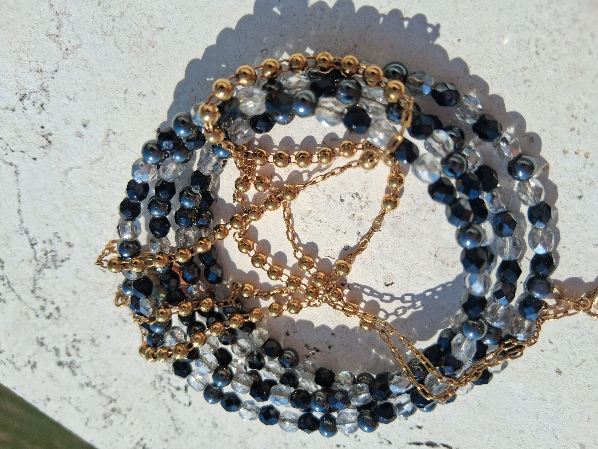 A close-up, overhead shot captures a tangle of beaded necklaces and chains arranged in a circular fashion on a textured, light-colored surface. The jewelry consists of clear faceted beads, opaque black faceted beads, and shiny dark gray or navy blue round beads, interspersed with small gold-colored ball beads and delicate gold chains. The arrangement is dense and layered, with some beads casting faint shadows on the surface beneath. The background is out of focus, showing hints of greenery in the upper left corner. The textured surface suggests it could be stone or concrete, possibly outdoors. The scene depicts a still life of accessory items, with no people or actions visible. The lighting suggests a sunny day, as indicated by the shadows. There is no discernible location within the city of Colorno, Italy.