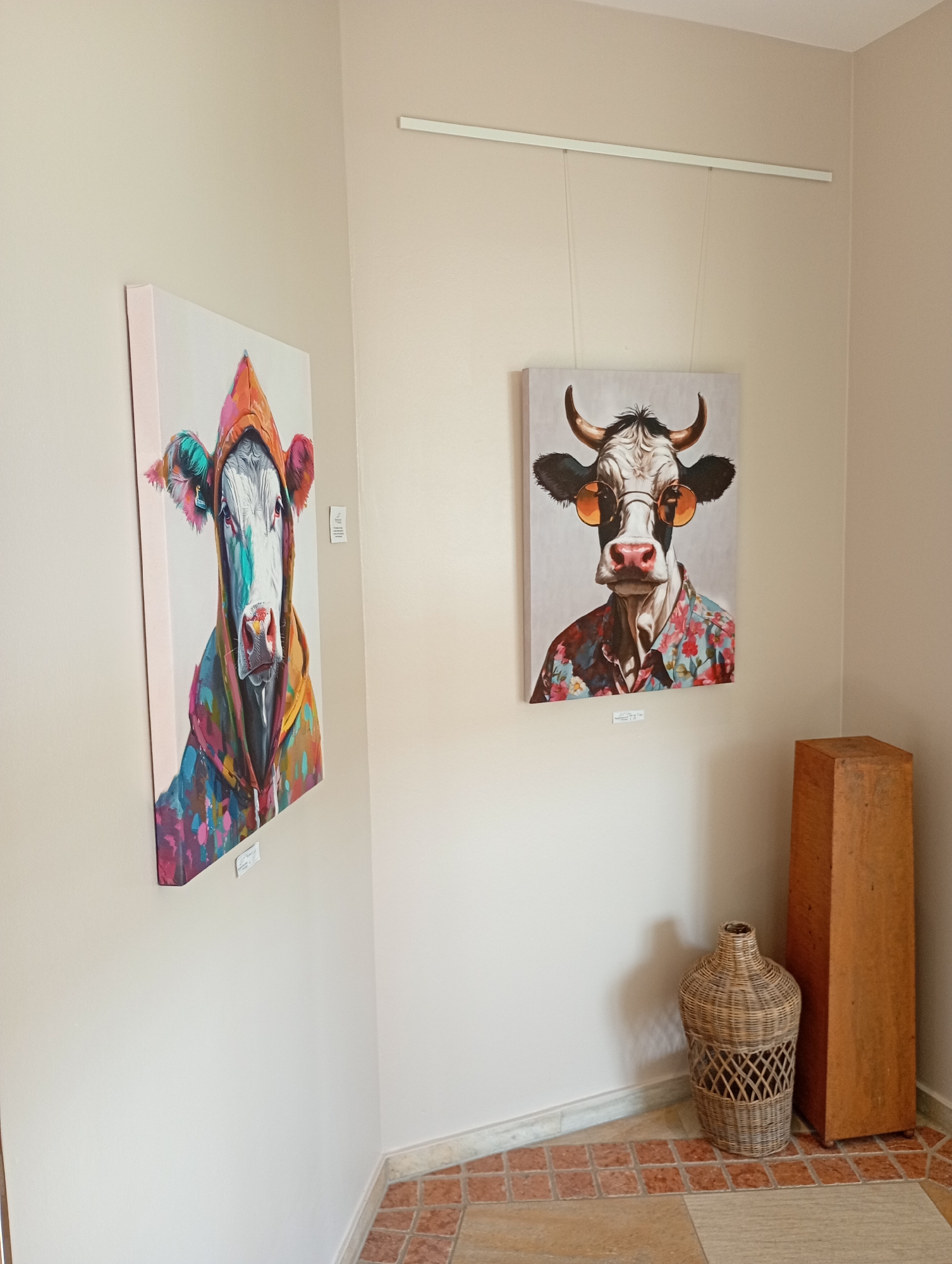 The image depicts an art exhibition corner in Bad Sachsa, Germany. Two framed paintings of cows are displayed on a light-colored wall. The painting on the left shows a cow wearing a colorful hooded jacket, while the painting on the right features a cow with sunglasses and a floral shirt. Beneath each painting, small white labels are affixed to the wall. To the right of the paintings, a woven basket and a tall, wooden rectangular object are placed on the floor, which is tiled with terracotta and beige squares. The lighting is neutral, illuminating the artworks and the surrounding space.