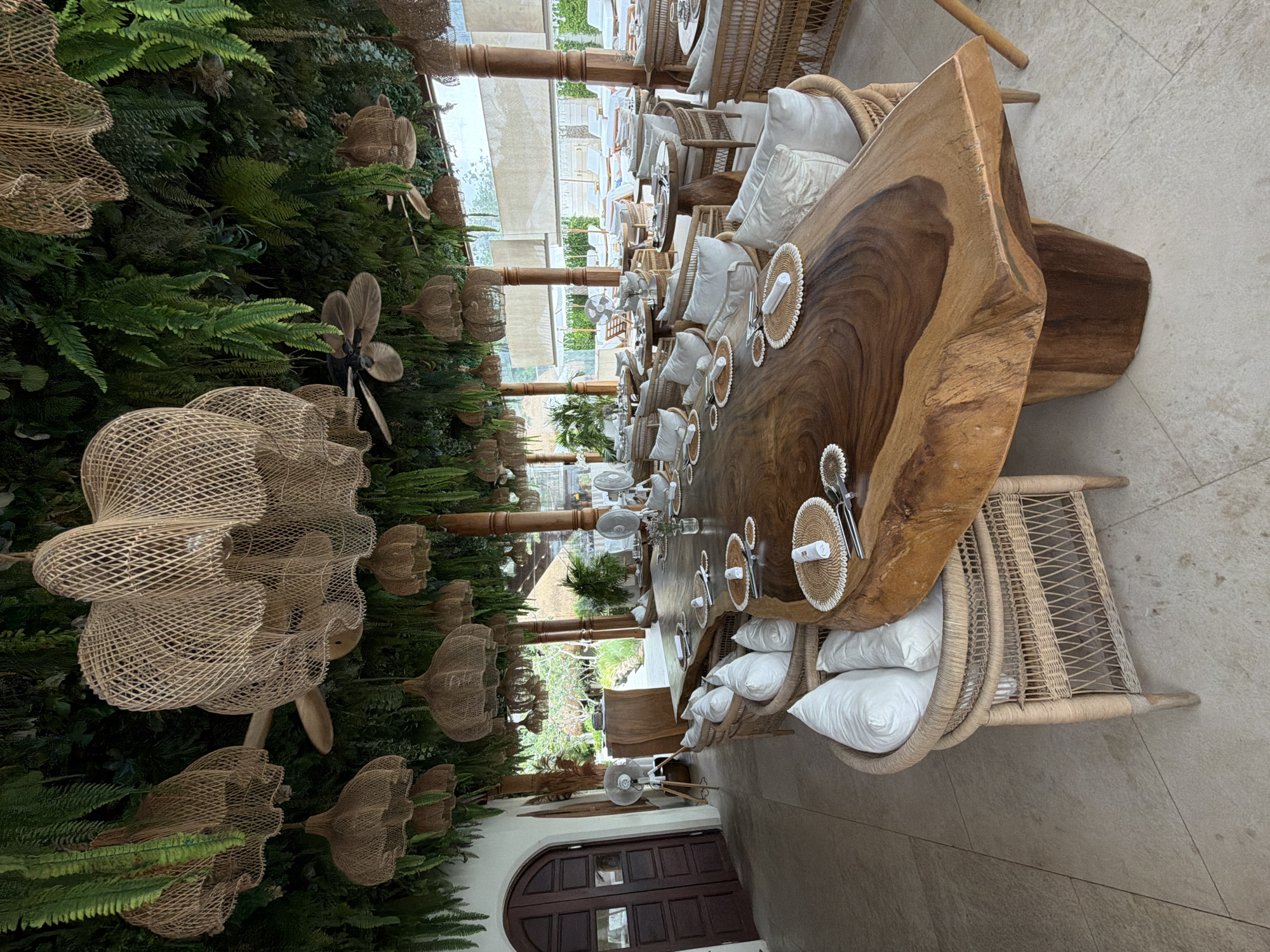 The image depicts a tropical outdoor dining area at a resort in Koh Samui District, Thailand. A large, live-edge wooden table is set with placemats, napkins, and cutlery, surrounded by wicker chairs adorned with white cushions. Overhead, a dense canopy of lush green foliage and woven rattan light fixtures create a natural, airy ambiance. Ceiling fans are visible amongst the greenery. In the background, additional tables and chairs are set up, and beyond that, lounge chairs and umbrellas suggest a poolside or beach area. The architecture features open-air structures with wooden pillars and light-colored flooring. The overall scene conveys a relaxed, upscale dining experience in a tropical setting.