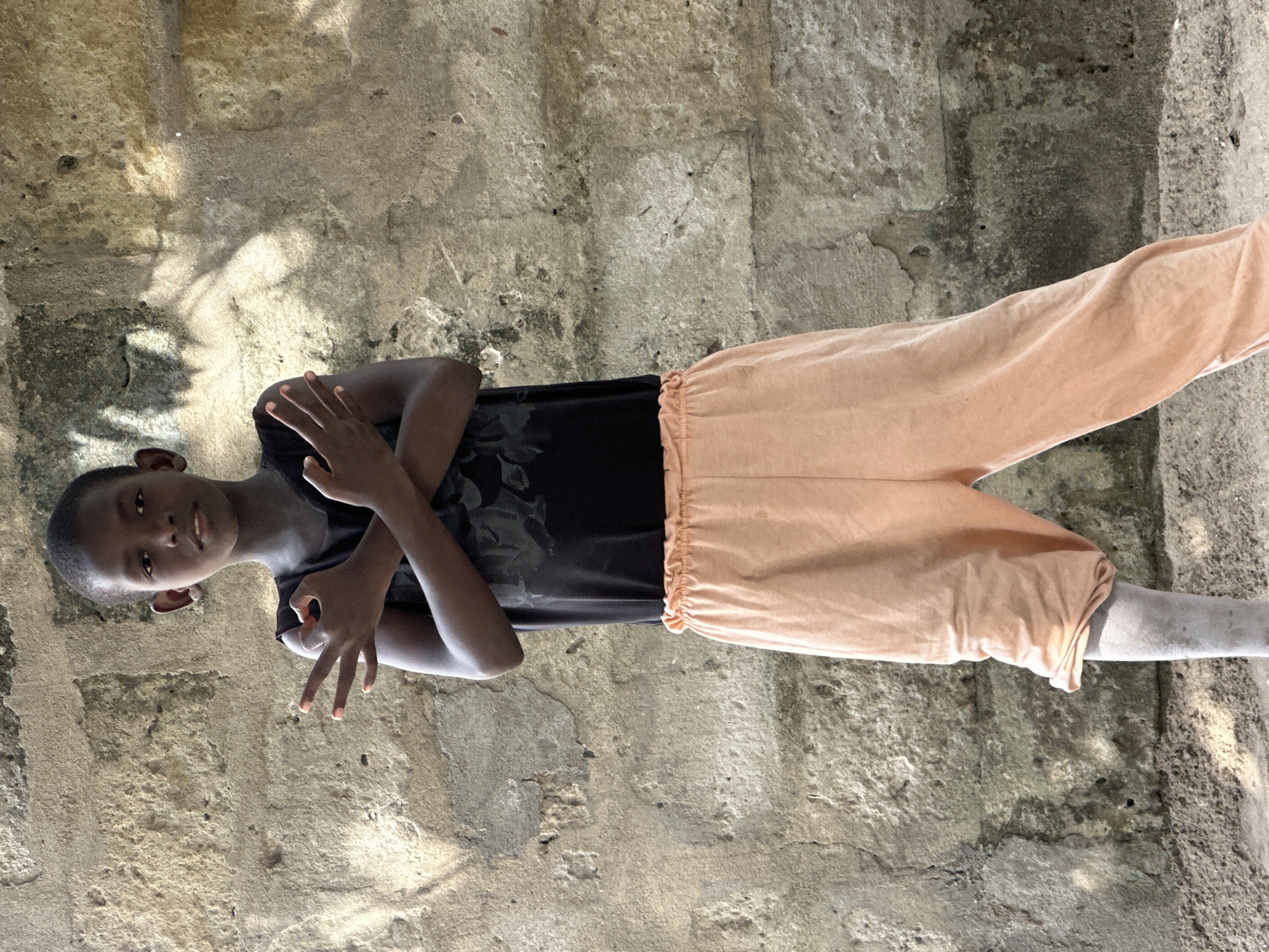 A young person stands in front of a textured, light-colored stone wall. The person is wearing a dark sleeveless shirt with a subtle floral pattern and peach-colored pants with a gathered waistband. Their arms are crossed at chest level, with their right hand forming a gesture. Their left leg is visible, appearing to have a lighter-colored, possibly gray, covering or is naturally lighter in tone. The ground is composed of sand and small stones. The overall lighting suggests an outdoor setting with dappled shadows cast on the wall, indicative of natural light. The scene appears to be in an outdoor environment, likely a residential area in Badagry, Nigeria, given the architectural style of the wall and the general setting.