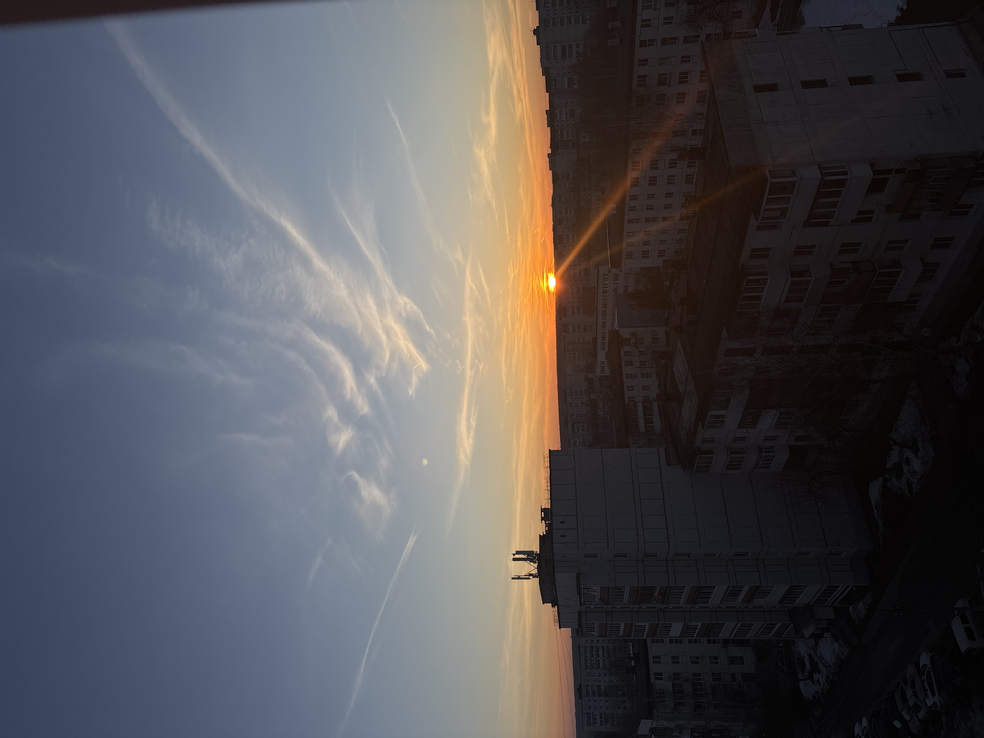 The image depicts a sunset over Chaikovsky, Russia. The sky transitions from blue to orange, with wispy clouds stretching across the frame. The sun is positioned on the horizon line, casting a bright glare and a golden hue over the cityscape. The urban environment includes rows of apartment buildings. The buildings are mostly gray and white. A few bare trees are visible. The photo is taken from an elevated perspective, possibly a higher floor in one of the buildings. The image is rotated 90 degrees.