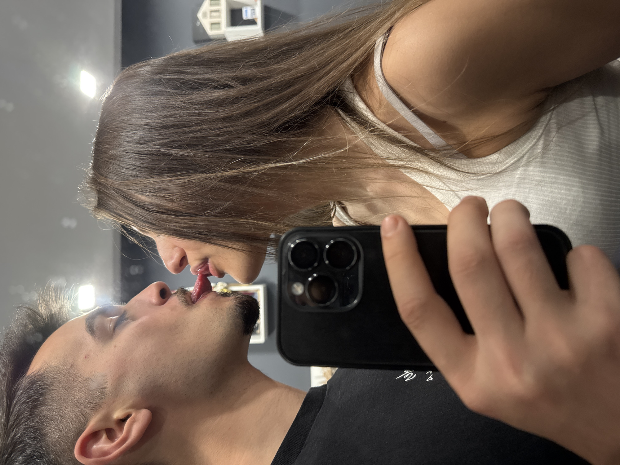 An indoor mirror selfie captures a playful and intimate moment between a woman and a man. The image is oriented vertically, with the woman positioned above the man.
The woman, on the upper left, has long, straight brown hair and is wearing a light-colored (possibly white or light grey) ribbed tank top with thin straps. The man, on the lower right, has short, dark hair, a mustache, and a beard, and is wearing a black t-shirt. Both individuals are engaged in a "tongue kiss" or a playful interaction, sticking their tongues out towards each other's mouths, almost touching.
A black smartphone, likely an iPhone due to its triple camera array, is held by a hand in the foreground, obscuring part of the man's chest and some background elements. The image appears to be a reflection captured by the phone in a mirror.
In the background, behind the woman's head, a bright, rectangular light source is visible. Further back, a small white decorative house model is perched on a surface. The rest of the background is blurred and out of focus, suggesting plain walls or surfaces in an indoor setting.
On the man's black t-shirt, some faint white stylized writing, possibly an unidentifiable logo or text, is partially visible. The scene suggests an affectionate and lighthearted interaction between the two individuals. The location is an indoor setting, and while the context states Sofia, Bulgaria, there are no specific visual cues within the image to confirm this.