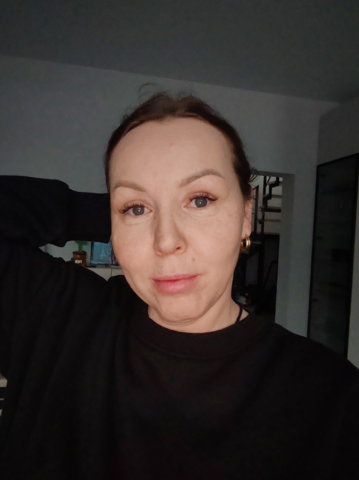 A woman with fair skin, light eyes, and dark hair pulled back poses indoors. She wears a black long-sleeved shirt and gold hoop earrings. Behind her is a glimpse of an interior: a doorway, stairs, and a television. The setting is not definitively recognizable as the city of Chaikovsky, Russia.