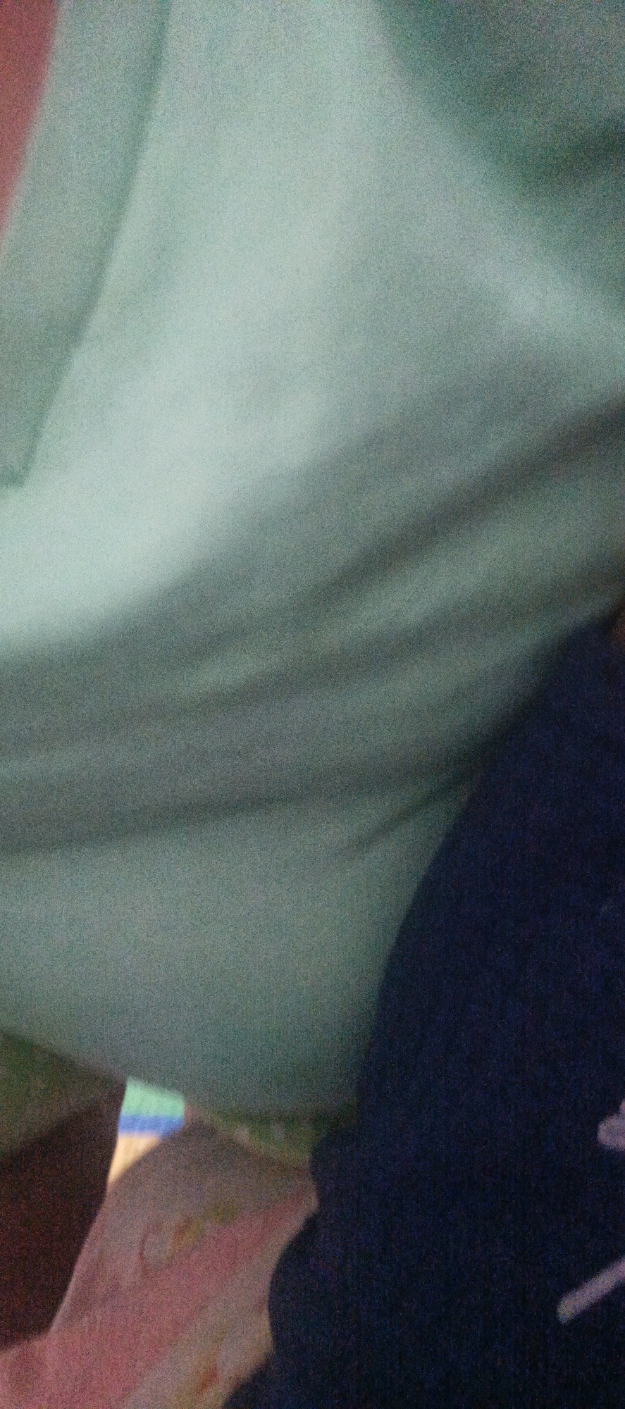 The image is a close-up, blurry, and grainy photograph of fabric. The dominant color is a muted teal, with darker navy blue fabric in the bottom right corner. There are subtle folds and shadows in the teal fabric, suggesting it is draped or wrinkled. In the bottom left quadrant, there appears to be a portion of pink fabric with some multicolored markings, possibly a patterned textile. The overall impression is of soft materials. The image does not contain any elements that would allow for the identification of a specific location in Cinta Dame, Indonesia, or any discernible people, subjects, actions, or interactions. The image is solely focused on the texture and color of the fabrics.