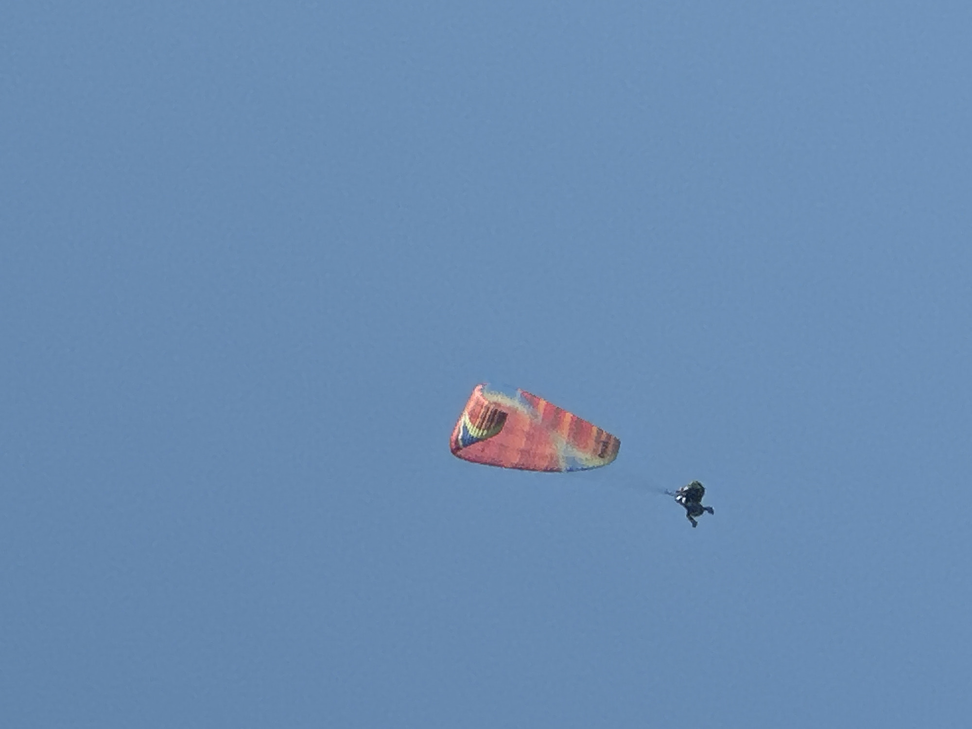 A paraglider with a multicolored wing, predominantly red, yellow, and blue, is captured in mid-flight against a clear blue sky. The paraglider is positioned in the upper left quadrant of the image. Below the paraglider, a person wearing dark clothing is suspended in a harness. The scene does not provide any specific landmarks or environmental context that would indicate its location within the city of Boreti, Montenegro. The focus is solely on the paraglider and the individual.