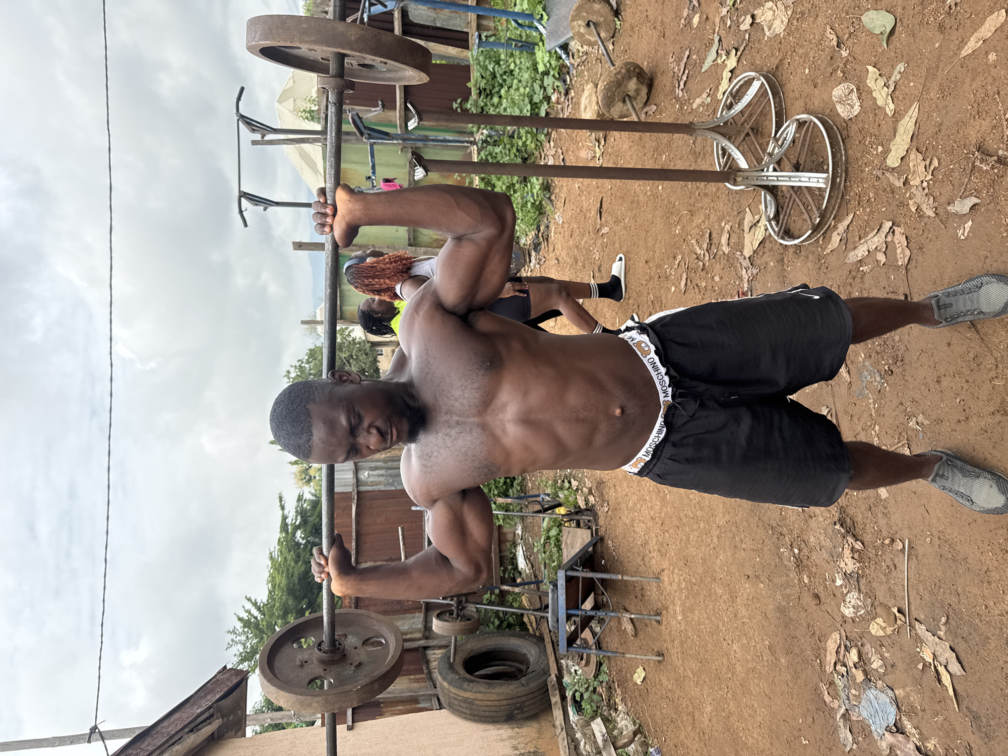 A shirtless Black male with dark skin, black hair, and a short beard is performing an overhead press with a barbell in an outdoor gym. He is wearing black athletic shorts with a white waistband that has a repeating bear graphic and the word "MOSCHINO". He is also wearing grey athletic shoes. The barbell has large, rusty metal weight plates. The gym appears to be rustic, set in a dirt-floored area with various pieces of makeshift exercise equipment, including a tire, a blue weight bench, and metal racks. In the background, two Black females, one wearing a yellow top and black shorts, and the other with auburn hair and a grey tank top, are visible. The environment suggests an urban or semi-urban setting in Akwanga, Nigeria, with buildings and vegetation visible in the background under an overcast sky.