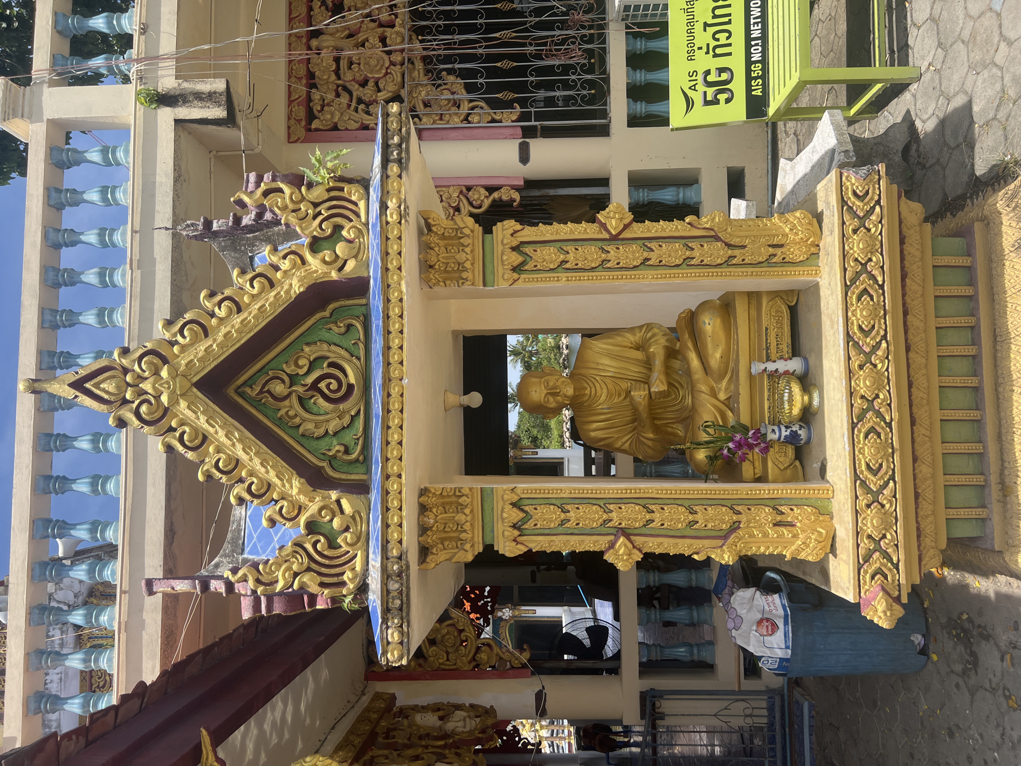 The image shows a golden statue of a seated monk within a richly decorated shrine. The shrine is ornate, featuring gold-colored carvings, green and red accents, and a tiered, roof-like structure adorned with intricate patterns. In front of the statue, there is a small vase with purple flowers, a decorative bowl, and another smaller vase.
The shrine is situated outdoors, with a paved ground surface. To the left of the shrine, a blue trash bin is visible, containing a white bag with some text. Behind the shrine, a building with a cream-colored facade and a balcony with blue balusters can be seen. A bright green bench with an AIS 5G advertisement is placed to the right of the shrine. The background suggests a tropical environment with trees and a body of water visible through an opening in the shrine. The sky is blue and partially cloudy.
This scene is located in a public area, likely a temple or a religious site, on the island of Ko Samui, Thailand, given the presence of Buddhist imagery and tropical surroundings.