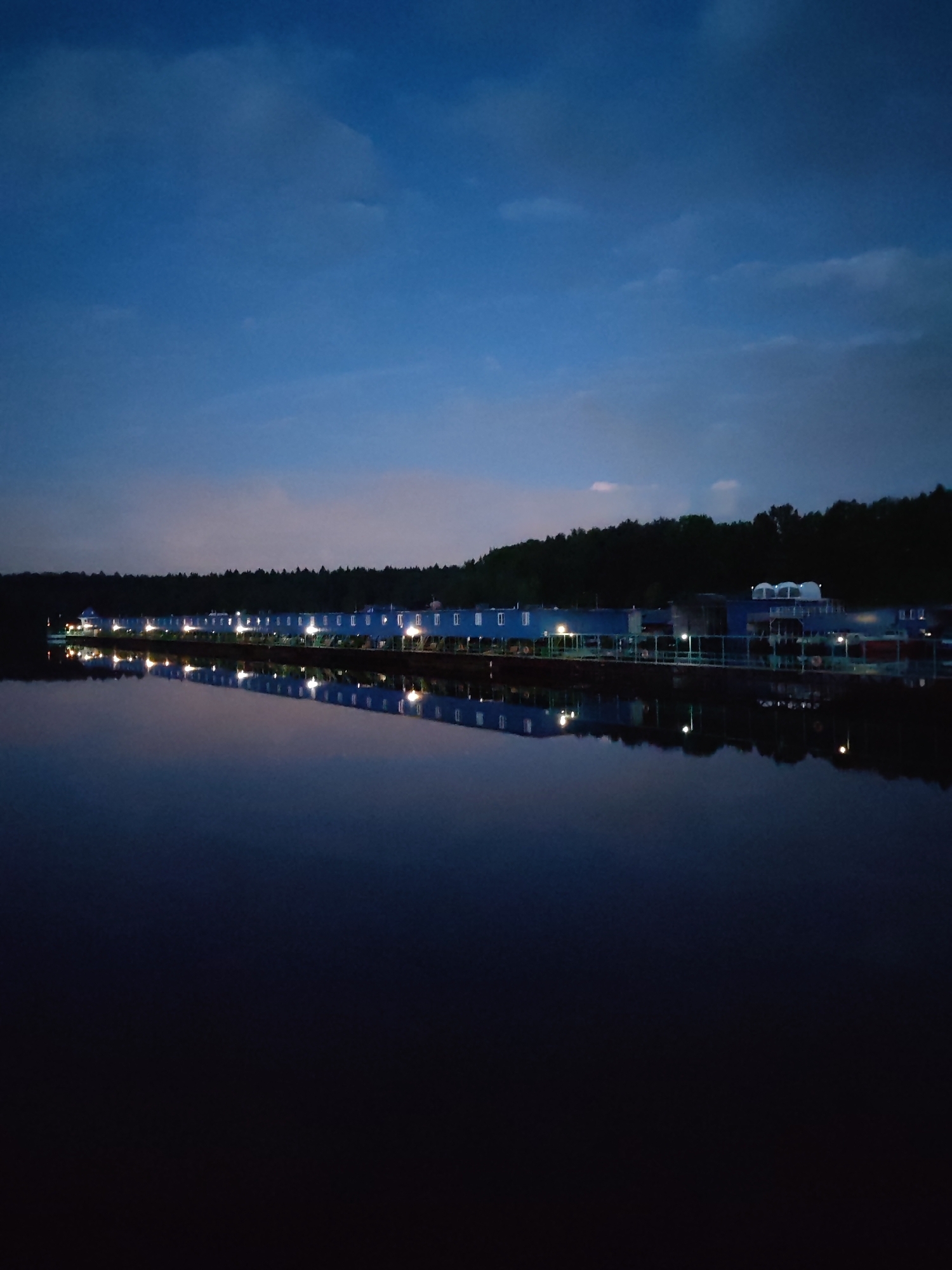 A nighttime shot features a body of water reflecting a row of buildings against a dark sky. The buildings are long and rectangular with evenly spaced windows, punctuated by intermittent lights, standing along the waterfront, adjacent to a densely wooded area. The sky is a mix of dark blue and hints of light clouds. The foreground consists of dark water and a smooth surface that mimics the buildings and lights along the shore. The location is Ermolino, Russia.