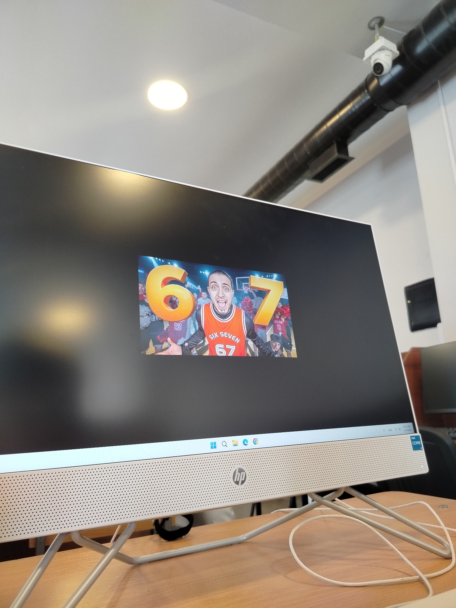 The image shows an HP all-in-one computer displaying a video featuring a man wearing a basketball jersey with the number "67" and the words "SIX SEVEN." The screen also has a large "6" and "7" next to the man. The computer sits on a wooden desk with its power cable visible.
In the background, a ceiling with a circular light fixture and a black pipe containing a security camera are visible. There's also a black rectangular object mounted on the wall.
It's not possible to determine the specific location in Yerevan, Armenia from the visual information provided.