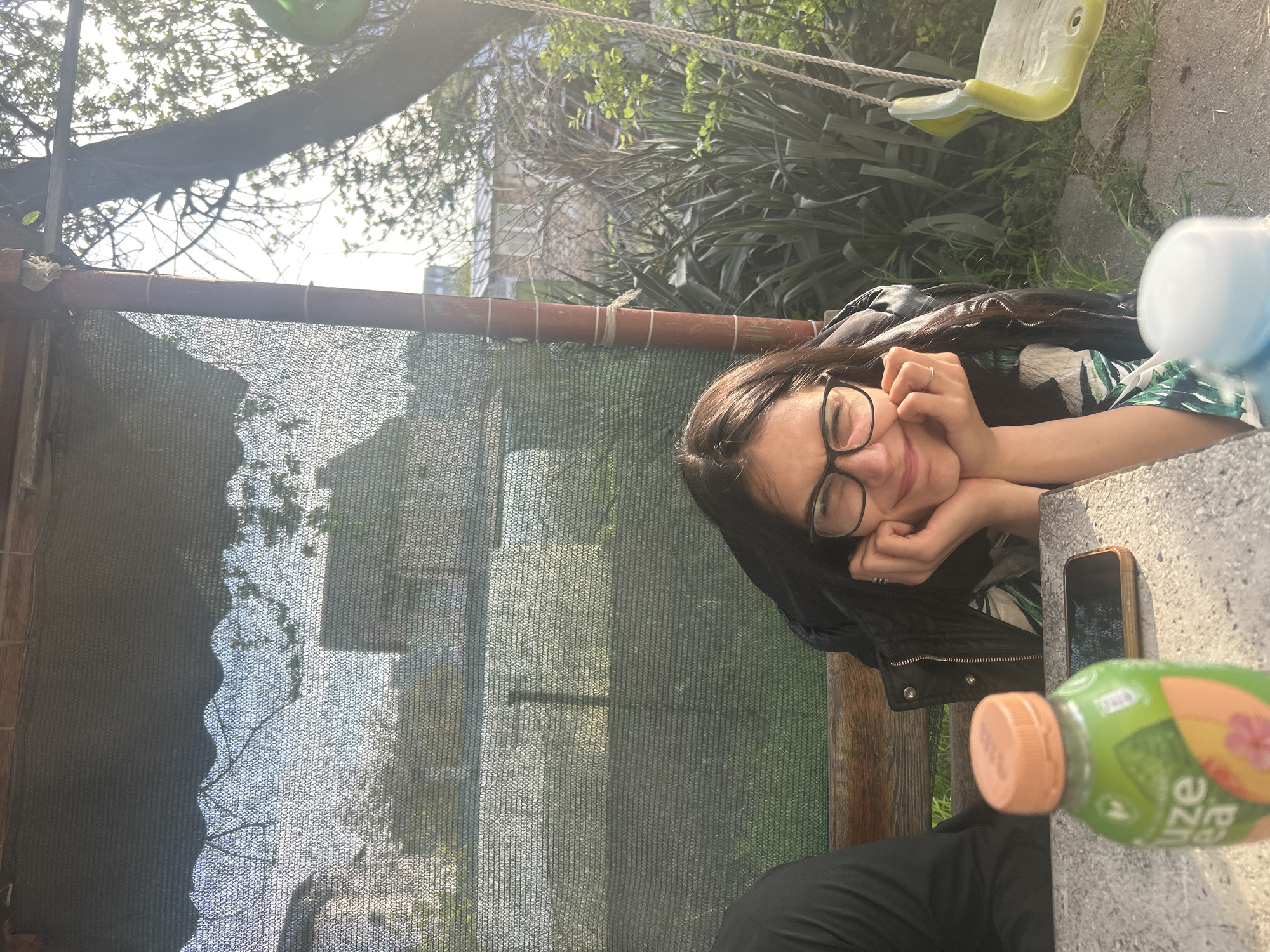 The image depicts a woman with dark hair and glasses smiling while resting her face on her hands at a table outdoors. A phone and a bottle of juice are on the table. Behind her is a fence-like structure with netting, and beyond that, trees and a glimpse of what appears to be urban structures. In the background there is also a child's swing. There are no clear signs indicating the specific location in Burgas, Bulgaria, but based on the urban backdrop and green surroundings, it may be a park or residential area in Burgas.
