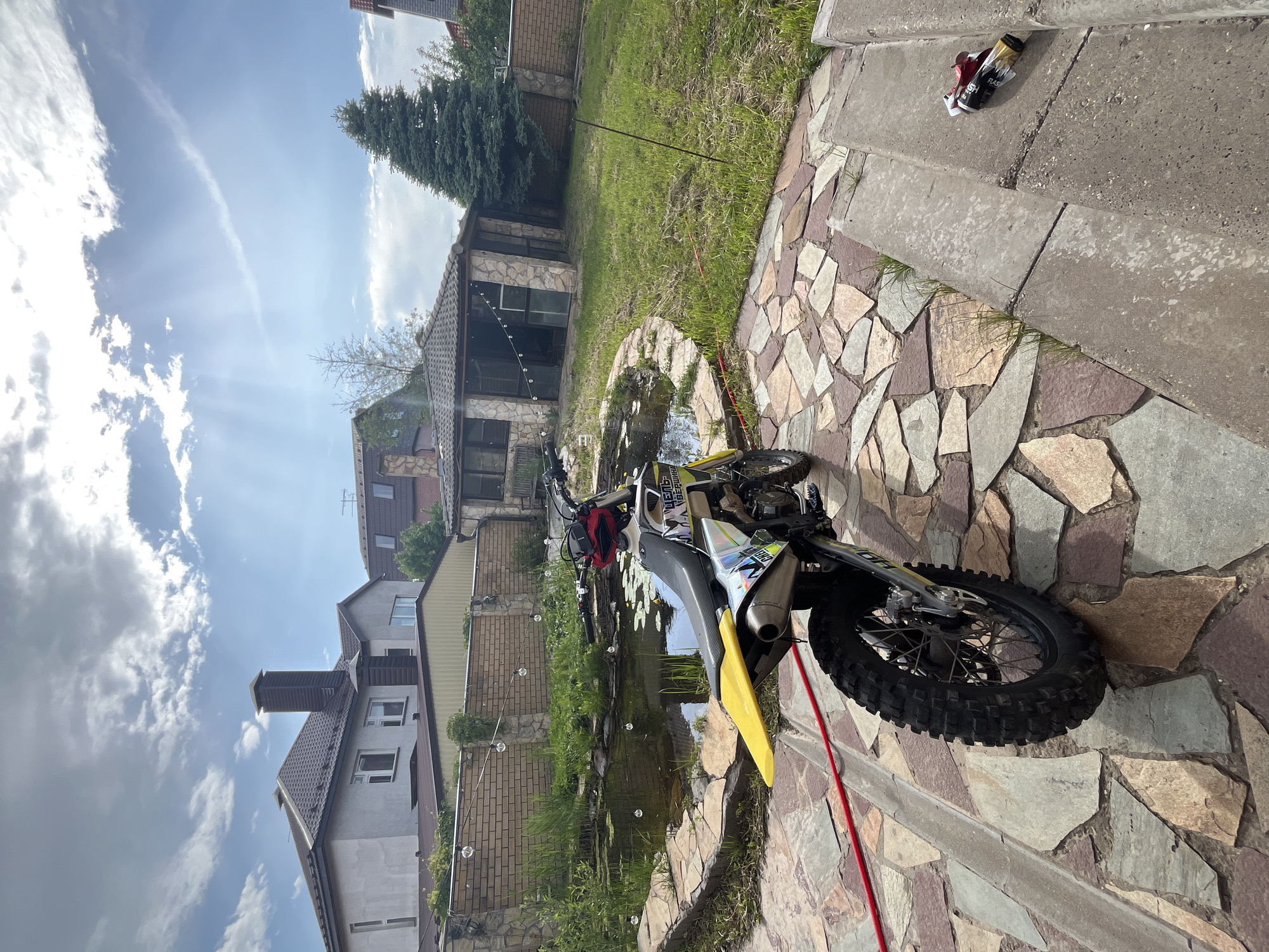 This media file captures a sunny daytime scene in a residential backyard in Ореховка, Russia. The image is composed of a stone-paved path in the foreground, leading towards a pond and garden area, with houses and trees forming the background.
The primary subject is a motocross-style dirt bike, parked on its kickstand on the stone path in the right foreground. It features a bright yellow front fender, black knobby tires, and colorful graphics on its frame, where parts of "HUSQVARNA" and numbers like "118" or "108" are visible. A red hose is laid out on the path near the bike.
The path itself is made of irregularly shaped, multi-colored flagstones. To the left of the path, a pond or water feature is visible, adorned with lily pads and numerous floating bubbles, suggesting a fountain or aerator. The pond is surrounded by green grass and lush vegetation. Beyond the pond and grass, a brick-built structure with large windows, possibly a gazebo or a smaller outbuilding, stands.
In the background, several houses with varied architectural styles are visible, some with stone facades and tiled roofs, and one with a prominent chimney. A tall evergreen tree stands to the right of one of the houses.
The sky is partly cloudy and bright, with visible sunbeams shining through the clouds, indicating good weather. On a concrete step in the upper right portion of the frame, several discarded items, including empty beverage cans, are visible. One can clearly shows the word "FLASH."
No people are present, and the scene appears static, showcasing the outdoor setting and parked vehicle.