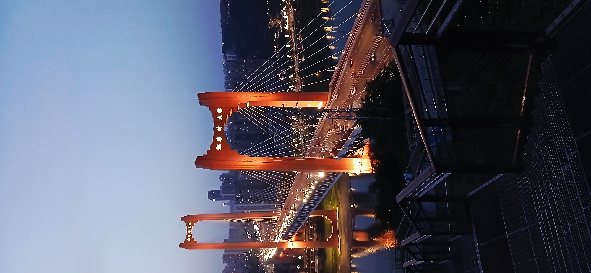 The image shows the Dongshuimen Bridge in Yuzhong District, Chongqing, China, at dusk. The bridge is a suspension bridge with orange towers. Cars move across the bridge. The scene is viewed from an elevated platform with metal railings. The city skyline is visible in the background. The river reflects the bridge's lights. The sky is a gradient of blue.