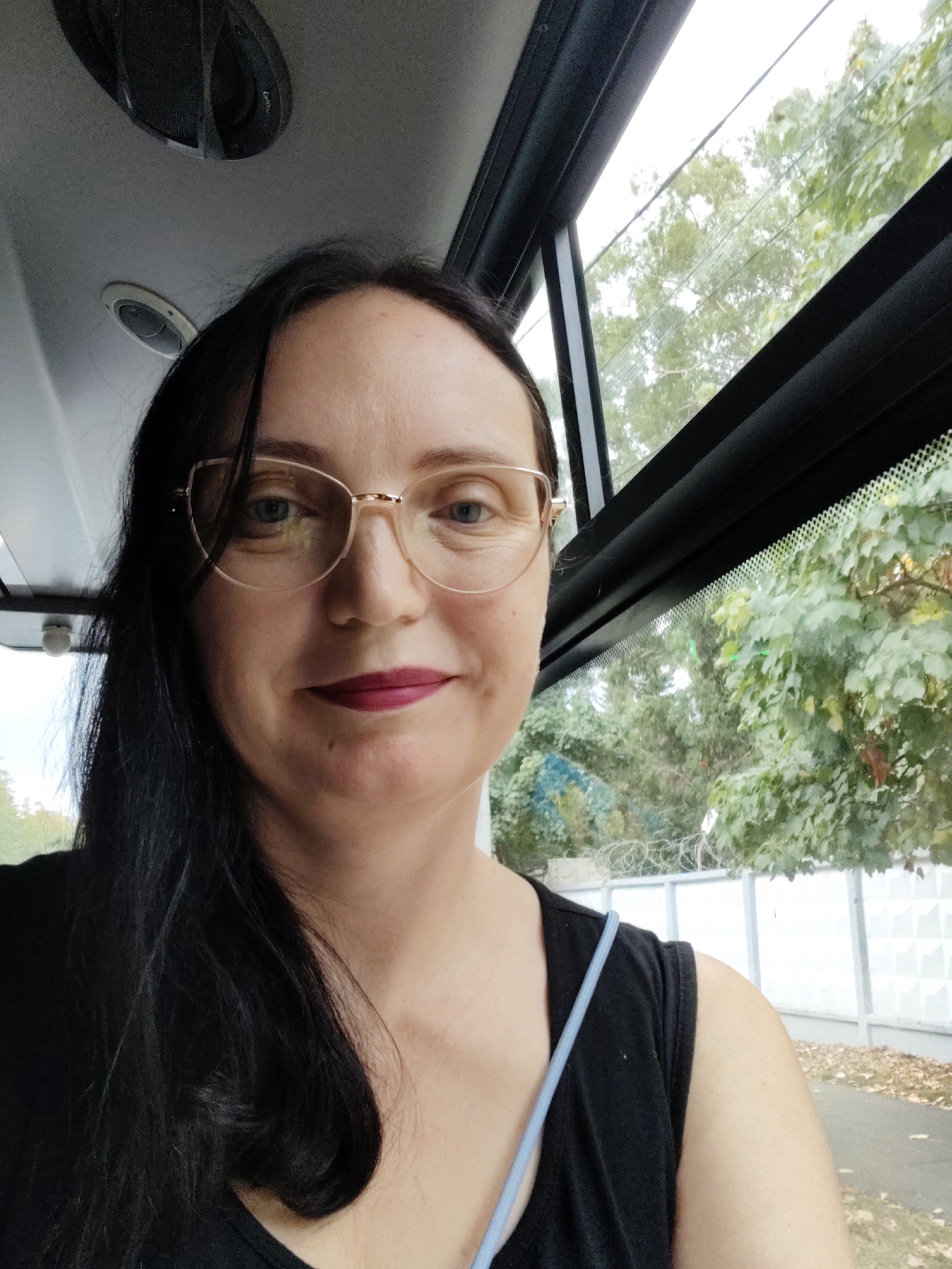 A medium shot photograph taken from inside a vehicle, likely a bus or van, shows a Caucasian woman with long, dark hair and glasses looking directly at the camera. She is wearing a dark top. Through the windows behind her, trees and a white fence are visible, suggesting an outdoor setting. The interior of the vehicle has a grey ceiling with ventilation grilles. The lighting appears to be natural daylight. The image does not contain enough information to determine the specific location within the city of Sochi, Russia.
