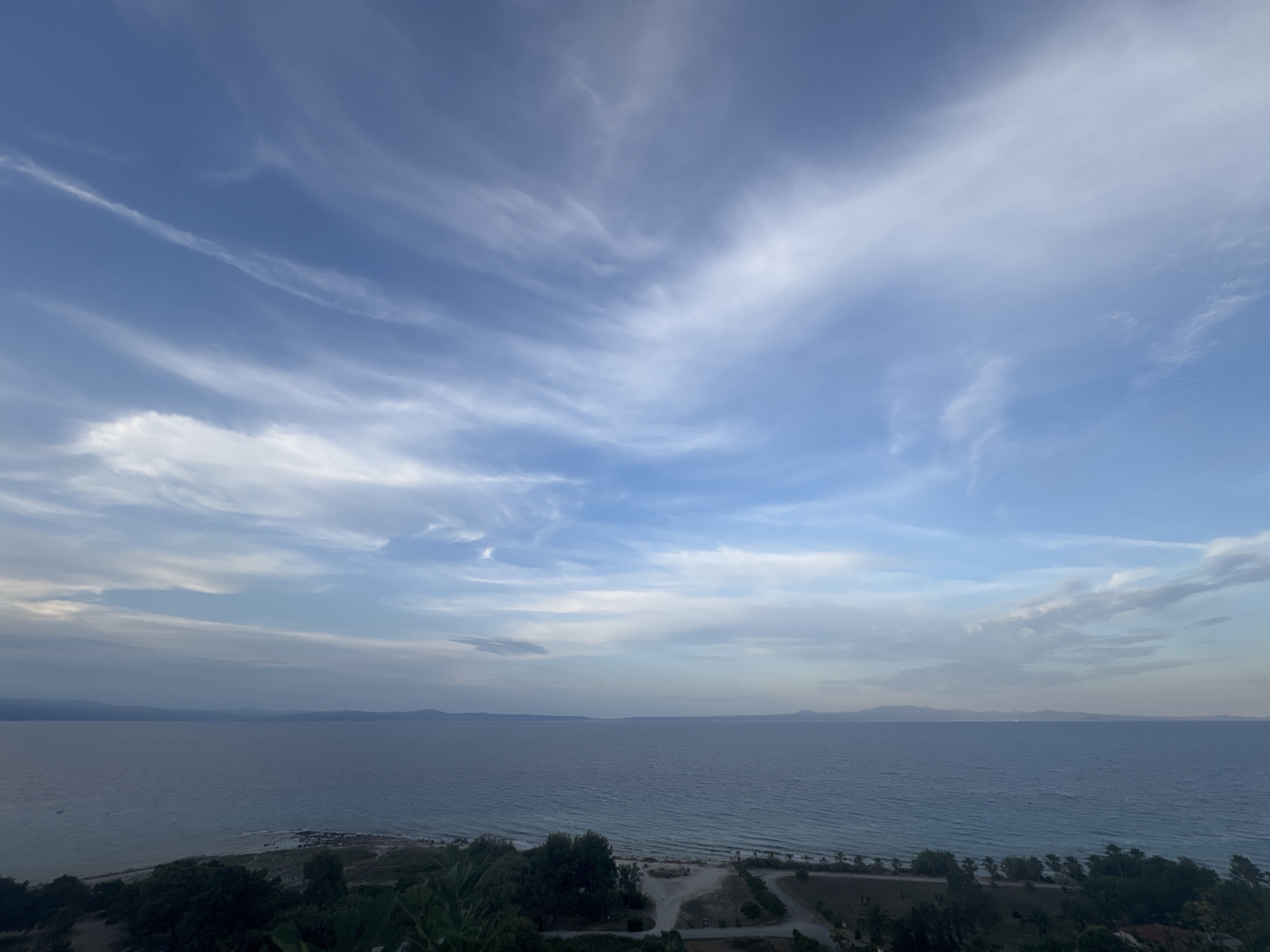 This is an eye-level, wide-angle shot of a tranquil seascape from a vantage point overlooking a coastline in Kassandra Municipal Unit, Greece. The sky is a dominant feature, filled with wispy, cirrus clouds painted across a vibrant blue expanse. The horizon is a soft blend of blues and grays, where the sea meets distant landmasses. The sea itself is a deep, calming blue, with subtle ripples indicating a gentle breeze. Along the coastline, a narrow strip of beach is visible, dotted with sparse vegetation and a few scattered trees. The foreground is partially obscured by the dark, lush foliage of trees, suggesting a natural or semi-developed landscape. There are no people or animals visible in the frame. The overall atmosphere is serene and peaceful, indicative of a coastal landscape during daylight hours.