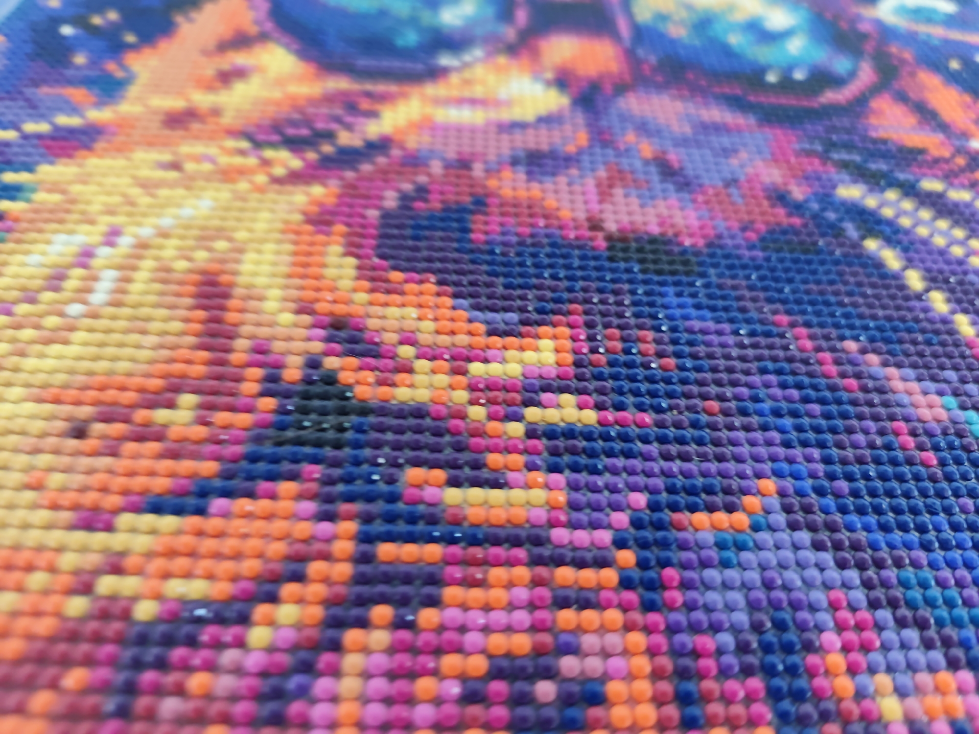 The image is a close-up, high-angle, shallow depth of field photograph of a diamond painting. The painting is composed of numerous small, round, colored resin beads arranged in a grid pattern to form a larger image. The colors visible include various shades of yellow, orange, pink, purple, and blue, suggesting a vibrant and possibly abstract or stylized subject matter. The texture of the beads is slightly uneven, with some appearing to catch the light. The focus is on the middle ground of the painting, with the foreground and background rendered out of focus. There are no people, animals, or identifiable landmarks visible in the image, and no specific location within Mykulychyn, Ukraine can be determined from the visual information provided. The scene depicts a craft or art project in progress or completed.
