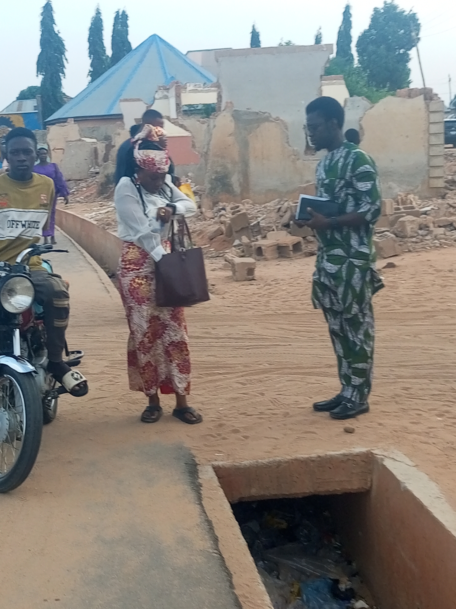 The image shows a man in a green patterned outfit standing and holding a book in front of a woman in a white blouse and floral skirt, who is holding a brown bag. To the left, a young man sits on a motorcycle wearing a mustard yellow "OFF WHITE" shirt. Behind them is a dilapidated building with a blue roof. The ground is sandy. In the foreground, there is a concrete drainage ditch filled with trash. The location appears to be a city, but the specific city and country are unknown.