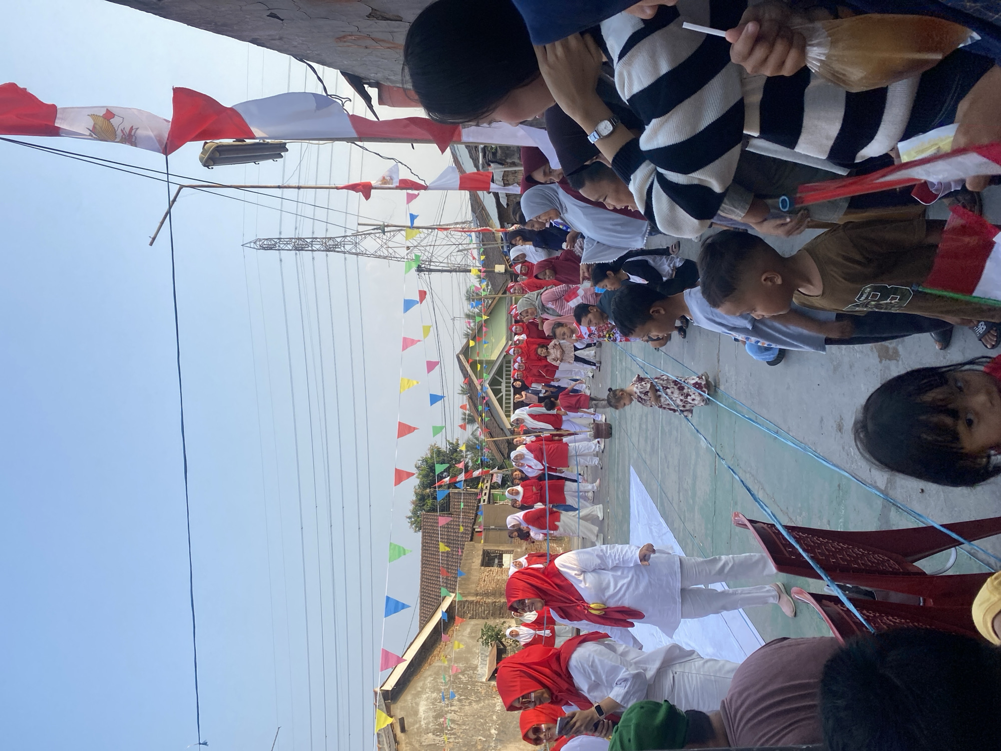 The image depicts an outdoor gathering in a residential area of Cilegon, Indonesia, likely for a local celebration or event. A group of people, many wearing red and white attire, are visible, suggesting a patriotic theme, possibly related to Indonesian Independence Day. Some individuals are dressed in hijabs. Several Indonesian flags are prominently displayed, including a large one on a pole. Decorative colorful triangular flags are strung across the street, adding to the festive atmosphere. In the background, residential buildings and a tall transmission tower are visible. Children are present, with some actively participating or observing. A few rows of red plastic chairs are set up, indicating an audience area. The scene is captured during daylight, with clear skies.