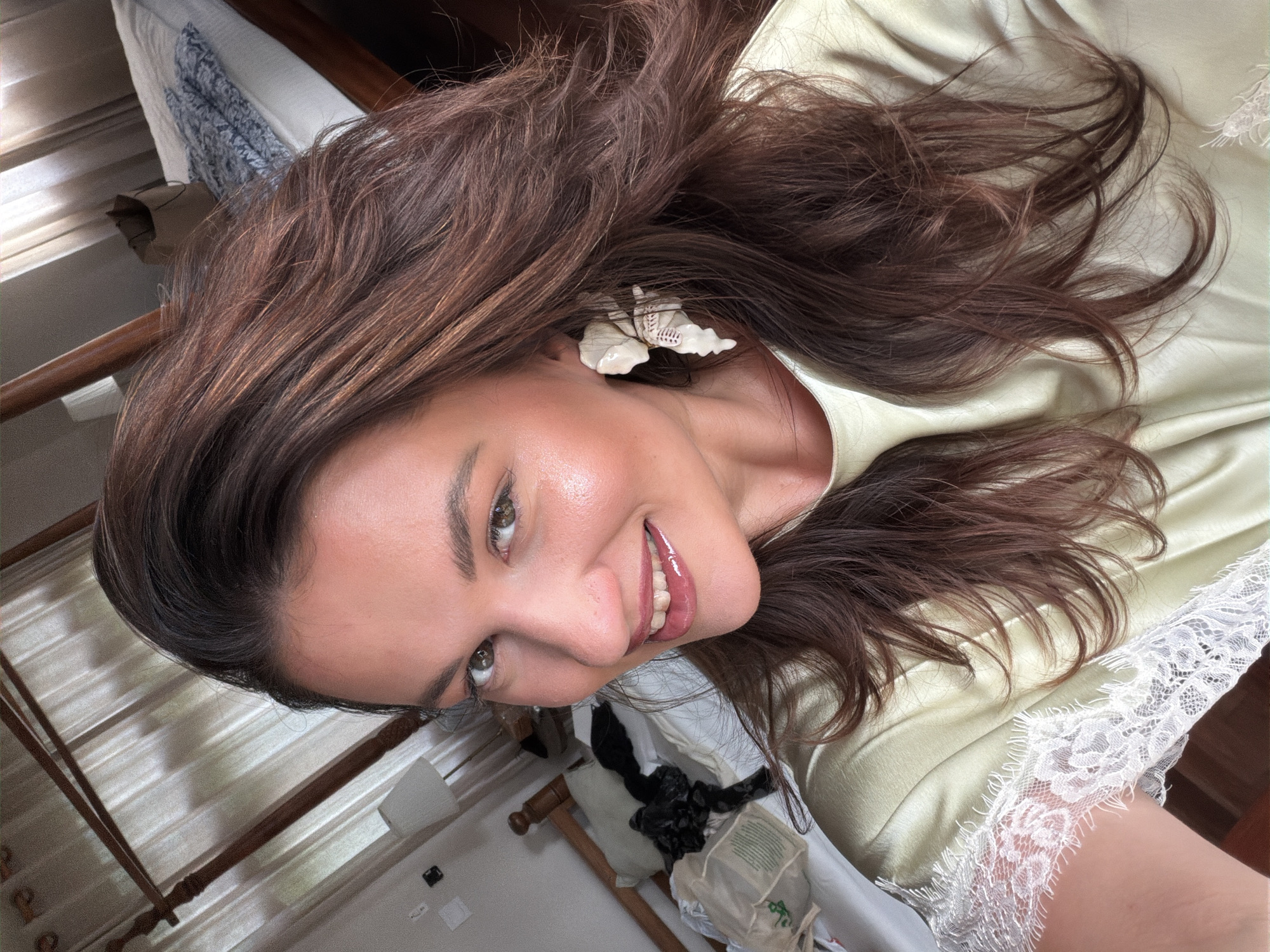 A woman with long, dark brown hair is looking into the camera, smiling. She is wearing a light green satin top with white lace trim on the sleeves. She has large white floral earrings. In the background, a four-poster bed with white linens and a wooden frame is visible. Curtains are drawn across a window behind the bed. A bedside table with a lamp and a paper bag are also present. The scene appears to be a bedroom in North Kuta, Indonesia.