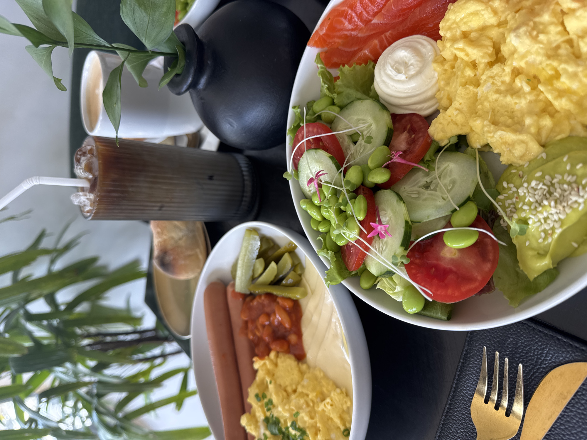 An overhead shot captures a visually appealing breakfast or brunch spread laid out on a dark tabletop. The scene features two main white bowls filled with various food items, along with drinks and decorative elements.
The larger bowl, positioned in the top right, holds a vibrant salad base of green lettuce, sliced red tomatoes, and sliced green cucumbers, generously dotted with bright green edamame beans, delicate white sprouts, and small pink edible flowers. Resting on this bed are distinct portions of thinly sliced smoked salmon, a swirl of white creamy dressing, a generous serving of fluffy yellow scrambled eggs, and half an avocado topped with white sesame seeds.
The smaller bowl, in the bottom left, contains two light brown sausages or hot dogs, a dollop of reddish-brown baked beans, several green pickled cucumber slices, and another portion of scrambled eggs, this time garnished with green chives or herbs. A pale yellow, possibly creamy or cheesy, substance is also visible next to the sausages.
Accompanying the meal are two drinks: a tall glass with a layered brown iced coffee or tea and a white straw, and a partially visible white ceramic mug containing a lighter-colored beverage. Decorative elements include a small black ceramic vase holding green leafy branches and a pair of golden-colored fork and knife resting on a black textured placemat. The overall presentation suggests a curated dining experience, possibly in a cafe or restaurant setting. No visible text is present.