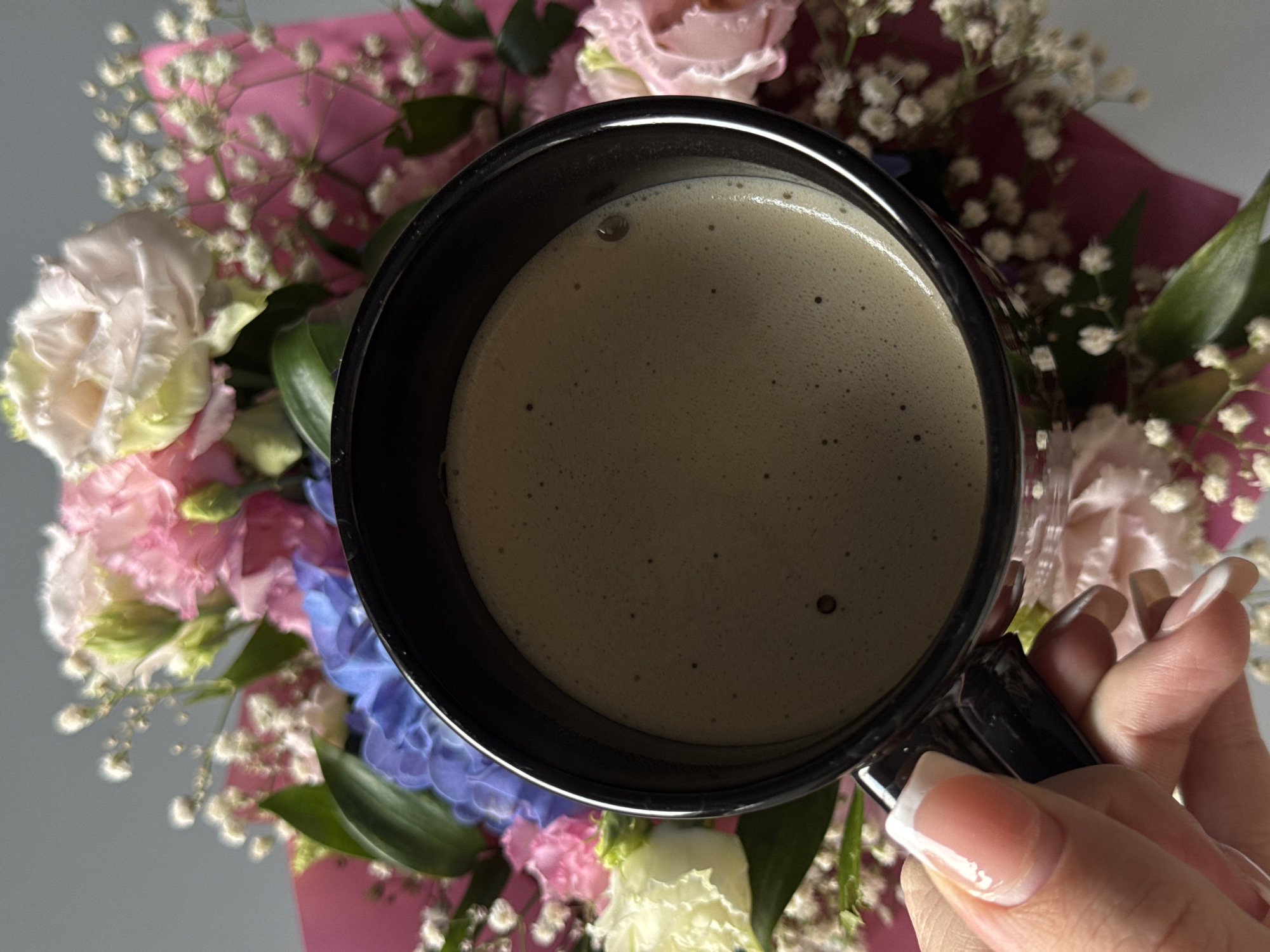 A close-up, top-down view shows a black mug of coffee with a frothy surface, held by a person's hand. The background is filled with a bouquet of flowers, primarily in shades of pink, blue, and white, with some green foliage. The lighting suggests an indoor setting. The image is a still shot and does not depict any specific location in Bratislava, Slovakia.