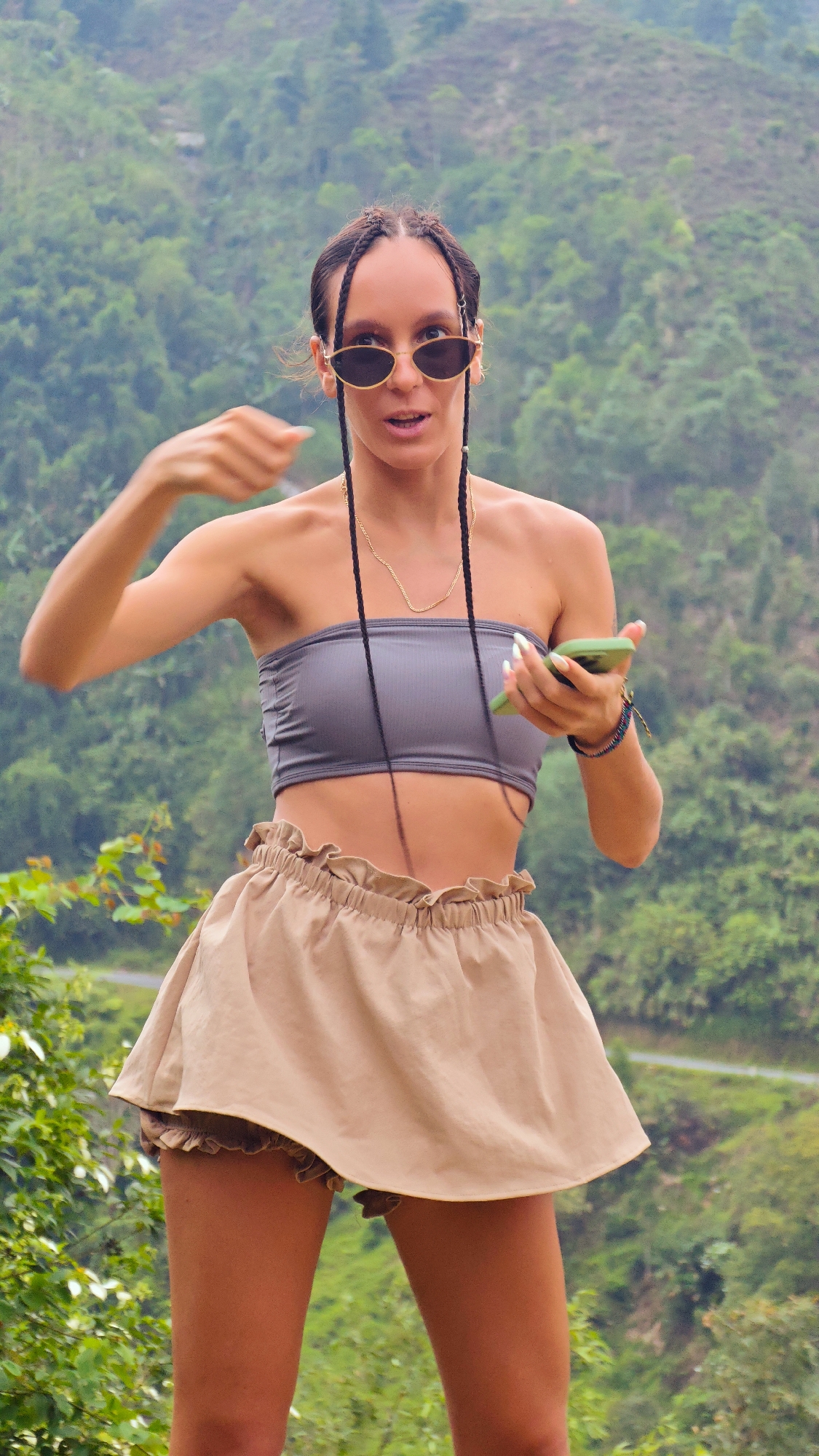 A young woman stands outdoors, captured in a full-body portrait against a lush green mountainous backdrop, indicative of Nậm Dịch Commune, Vietnam. She appears to be in her 20s or 30s, with dark hair styled in multiple long braids, some of which fall forward. Her attire includes a grey ribbed strapless bandeau top, light brown or beige high-waisted ruffled shorts that resemble a skirt, and oval-shaped sunglasses with gold frames. A delicate gold chain necklace hangs around her neck.
She is actively engaged, holding a green smartphone in her right hand while her left arm is raised with fingers slightly curled, suggesting a gesture, possibly pointing or emphasizing something. Her mouth is slightly open, conveying an impression of speech, surprise, or animated reaction. Several colorful bracelets adorn her right wrist, and her nails are manicured with a light polish.
The scene is set during daytime, with clear, natural lighting illuminating the dense foliage and rolling hills in the background. A faint road or path can be discerned winding through the distant mountains. The overall setting is a vibrant, natural landscape. No text is visible in the image.