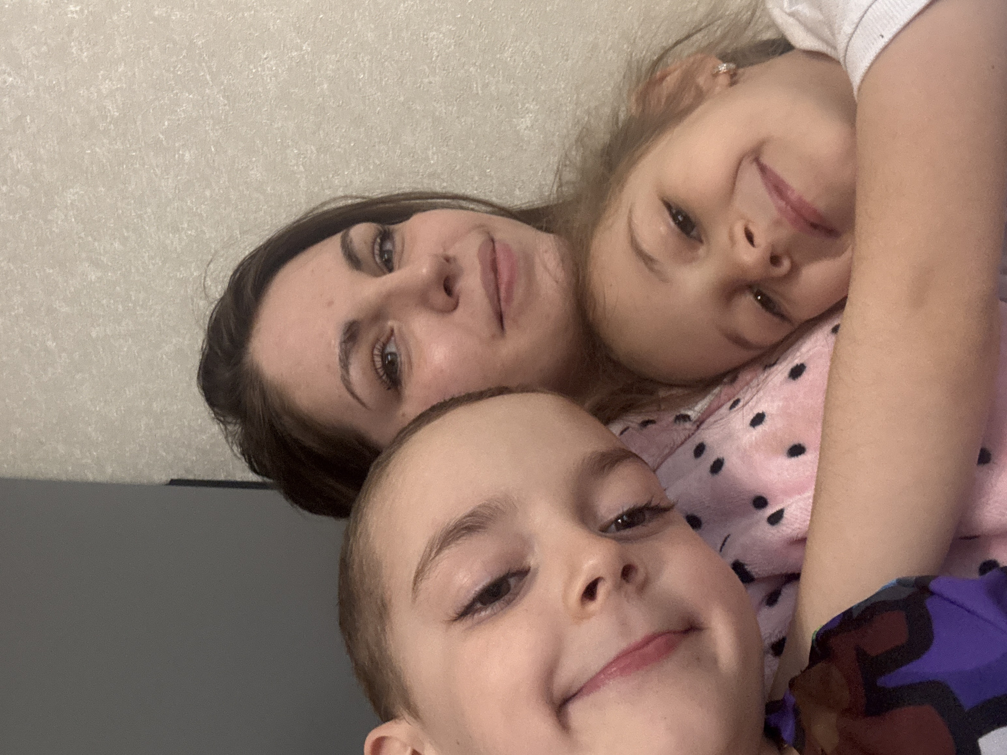 A selfie captures a woman and two children in close proximity. The location cannot be determined with the provided information. The background appears to be a wall, possibly indoors. The subjects are smiling and appearing to interact affectionately. One child wears a pink shirt with black polka dots. The other child wears a purple patterned shirt.