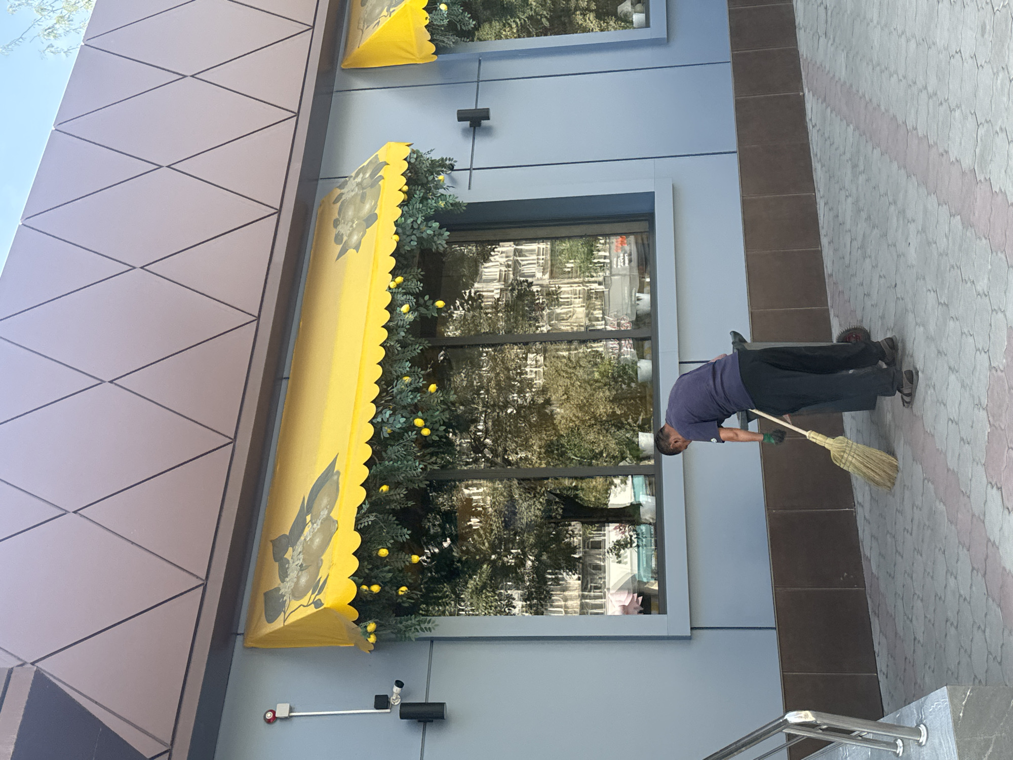 A man is sweeping a patterned pavement in front of a building with a blue facade. The man is wearing a dark blue t-shirt, dark pants, and sandals. He is holding a broom and appears to be pushing a small cart with a wheel. Above him, a bright yellow awning is decorated with artificial greenery and small yellow orbs, possibly representing lemons. The awning has a patterned edge and a depiction of lemons on its surface. Behind the awning are large windows reflecting trees and buildings. The building's upper facade is covered with dark metallic panels arranged in a diamond pattern. The scene appears to be outdoors, possibly on a street or walkway in a city. This location is identified as Bishkek City, Kyrgyzstan.