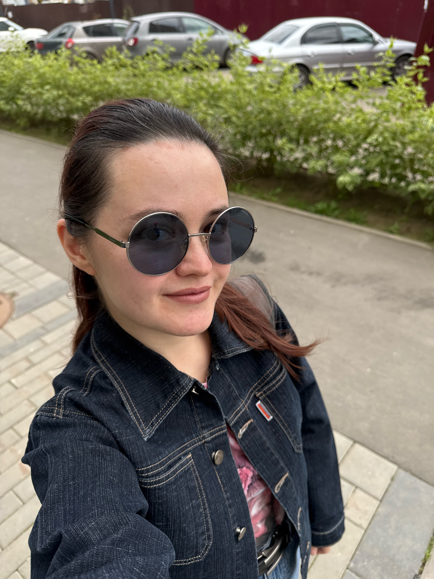 A medium shot photograph taken from a high angle depicts a young woman in a denim jacket and sunglasses, looking towards the camera. She has dark hair pulled back and is smiling faintly. Behind her, a row of green bushes is visible, with cars and a building in the background, suggesting an urban or suburban environment. The ground is paved with light-colored tiles. The scene appears to be outdoors, possibly in a residential area or a park in Izhevsk, Russia, given the context.