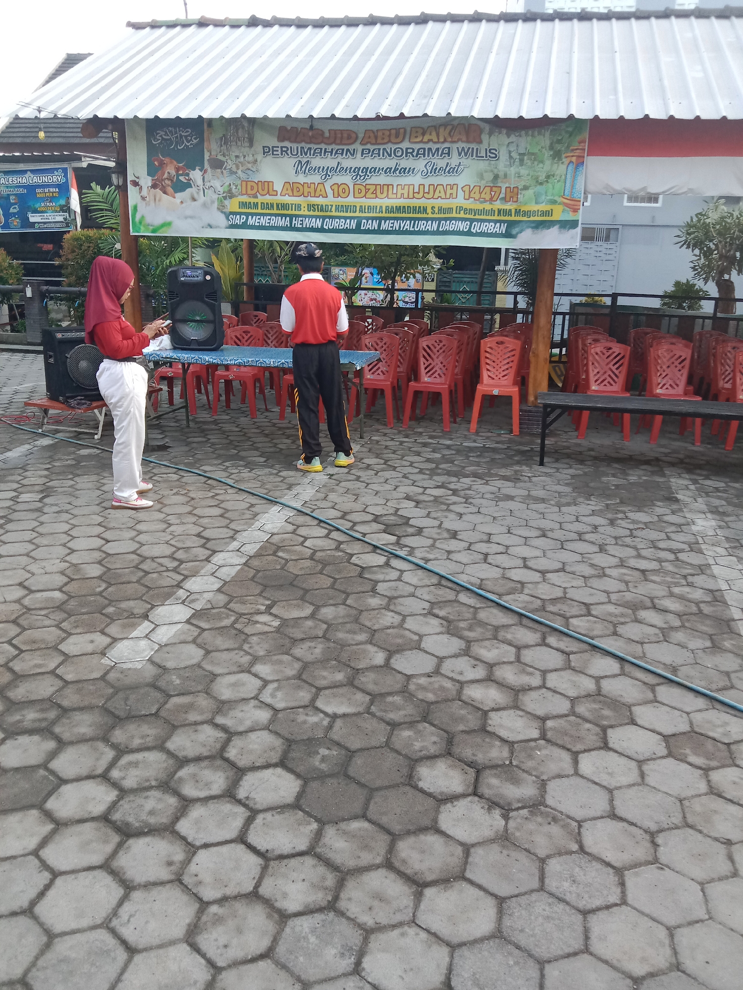 The image depicts an outdoor scene in Madiun, Indonesia, likely at a mosque or community gathering area. In the foreground, a person wearing a red hijab and white pants stands by a portable speaker, looking at a phone. Another person in a red shirt and black pants with yellow stripes is also present. Behind them, rows of red plastic chairs are set up, some on tables covered with blue cloths. A banner in the background announces an "Idul Adha 10 Dzulhijjah 1447 H" event at "MASJID ABU BAKAR PERUMAHAN PANORAMA WILIS," indicating the celebration of Eid al-Adha and offering of qurban (animal sacrifice). The ground is paved with hexagonal concrete tiles. The overall ambiance suggests preparation for a religious event or community function.