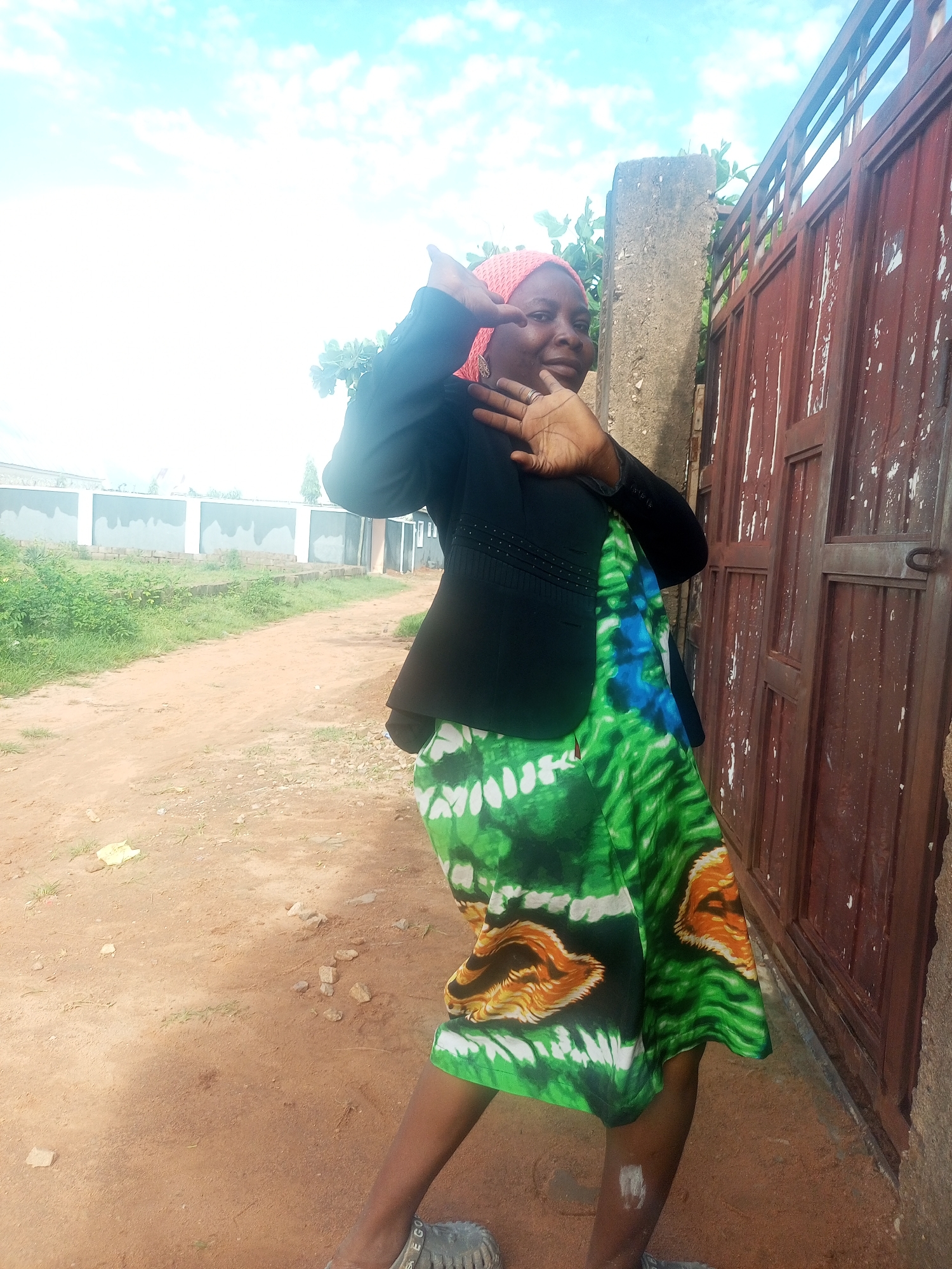A medium shot captures a woman in Tempure, Nigeria, posed in front of a rustic, weathered red metal gate. The woman, who appears to be in her late 20s or early 30s, is wearing a black blazer over a vibrant green and blue tie-dye dress. She has on a pink headscarf and is smiling at the camera with her left hand raised to her forehead, fingers spread. Her right hand is extended outwards, palm facing the viewer. The background reveals a dusty, unpaved road leading to what appears to be a construction site with a low concrete wall and some buildings under construction. Green vegetation flanks the road, contrasting with the dry, earthy terrain. The sky above is bright blue with scattered white clouds.