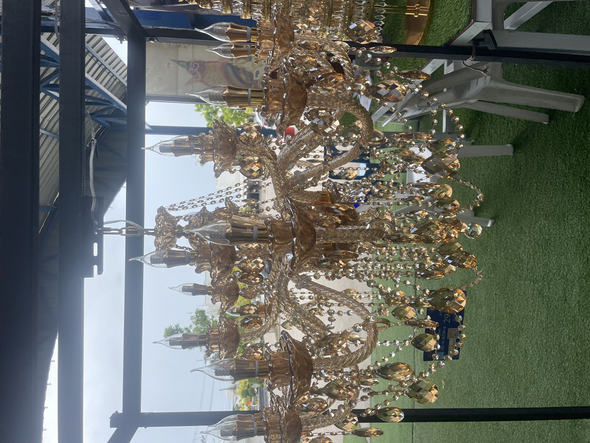 A large, ornate, gold-colored chandelier with numerous amber-toned crystals hangs from a dark metal frame. The chandelier features multiple tiers and arms, each holding a candle-shaped lightbulb. The crystals are strung together with smaller clear beads, creating a cascading effect. The chandelier is positioned in front of a window or open space, through which a glimpse of the outdoors can be seen. The background consists of a green, artificial turf-like surface, white tables and chairs, and some greenery and buildings, suggesting an outdoor seating area or market. A blue price tag is visible on one of the crystal strands. The lighting is dim, and the overall impression is one of opulence and a somewhat dated aesthetic.