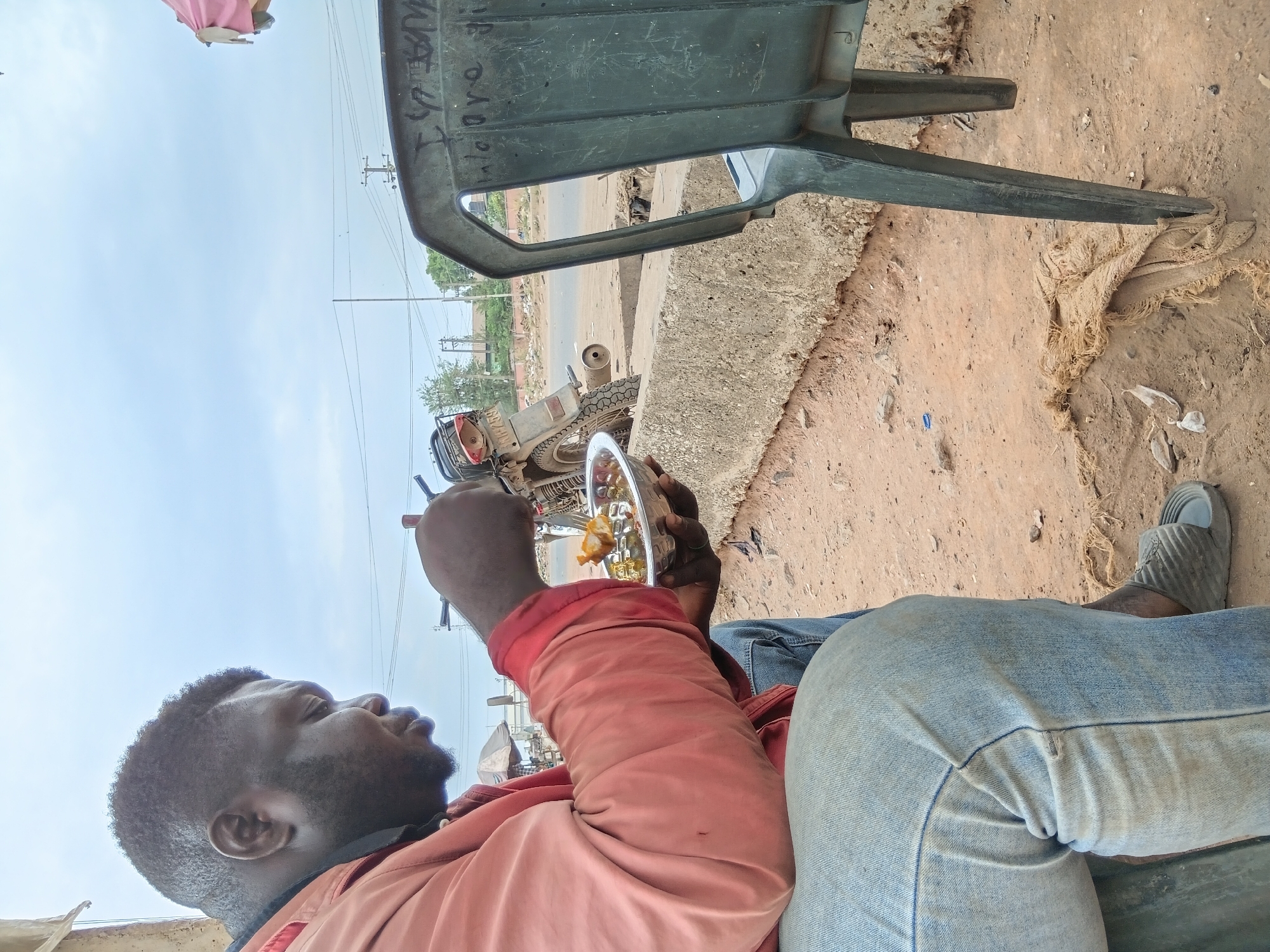 The image depicts a man sitting on a green plastic chair by the side of what appears to be a road in Akute, Nigeria. He is wearing a red shirt and blue jeans, and is eating from a plate. A motorcycle is parked nearby. The ground is dirt and sand. Overhead power lines and poles are visible in the background. The scene is captured from a low angle, slightly rotated.