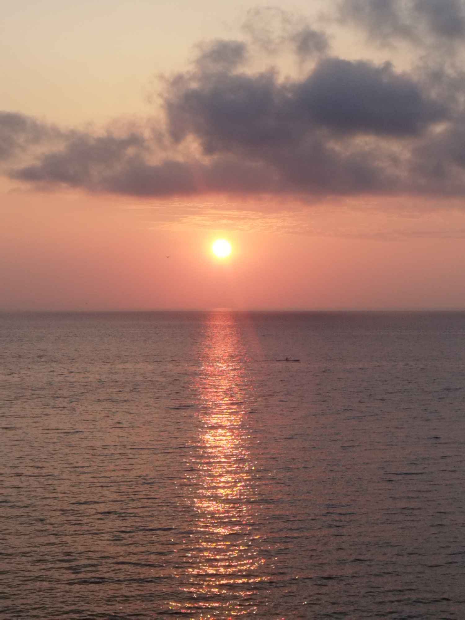 A wide-angle, vertical, eye-level shot captures a serene sunset over the ocean. The sun, a bright, diffused orb of light, is positioned slightly to the left of the frame's center, casting a luminous reflection on the water. Wispy, dark clouds drift across the upper portion of the sky, tinged with the warm hues of the setting sun. The ocean, a deep, shimmering expanse, exhibits gentle ripples, reflecting the sky's colors and the sun's glow. A small, dark boat, occupied by a person, is visible in the mid-distance, its silhouette a stark contrast against the bright water. The overall atmosphere is one of tranquility and natural beauty. This scene is consistent with a view from the coastal areas of Antibes, France, known for its picturesque sunsets over the Mediterranean Sea.
