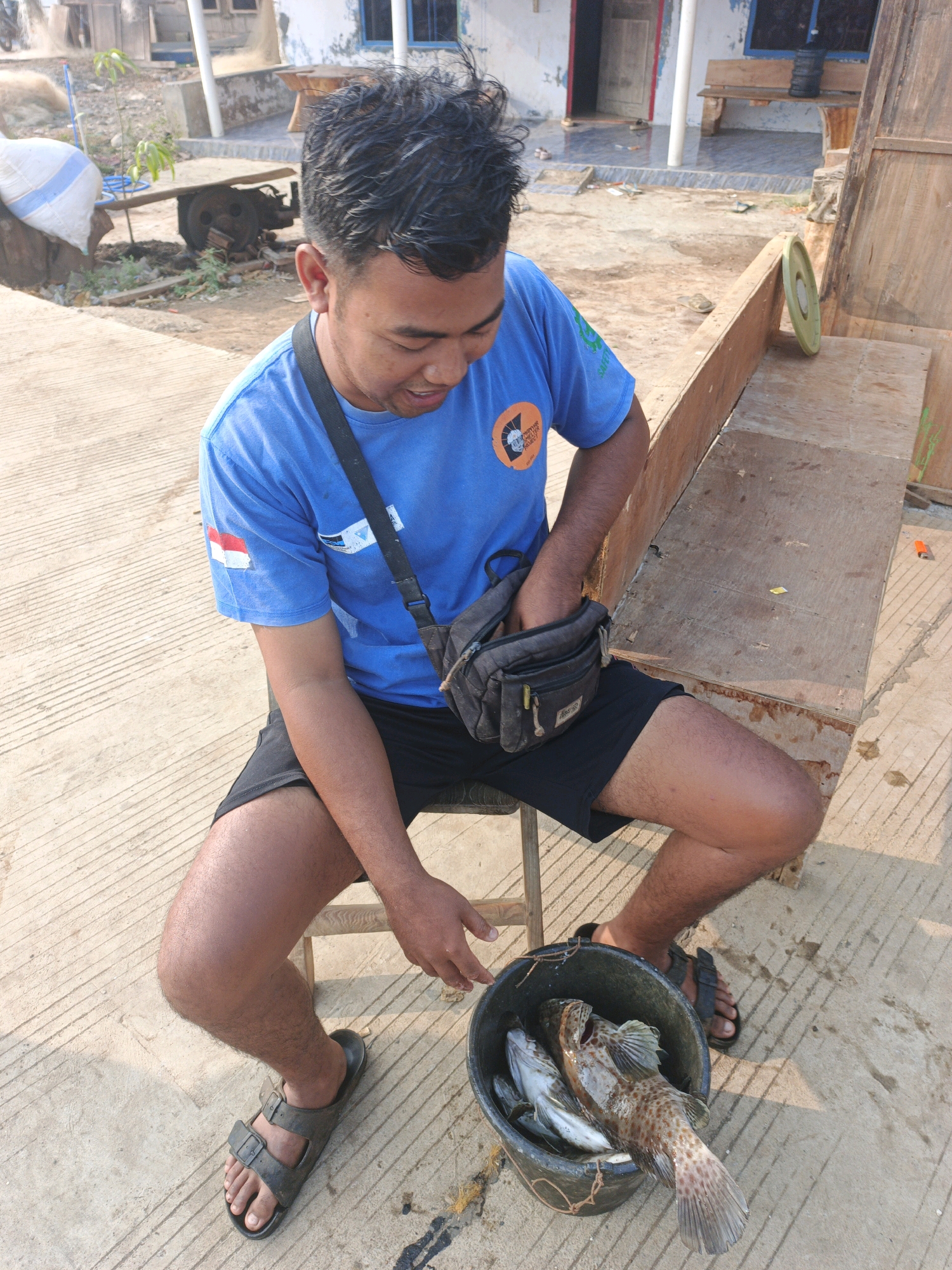 A man wearing a blue t-shirt and black shorts sits on a stool outdoors. He has a dark crossbody bag slung over his shoulder and is reaching into it. In front of him, on the ground, is a bucket containing three fish. The man is wearing black sandals. The ground appears to be concrete with some dirt and debris. In the background, there is a wooden structure and a building. The scene appears to be in a rural or coastal area in Indonesia.