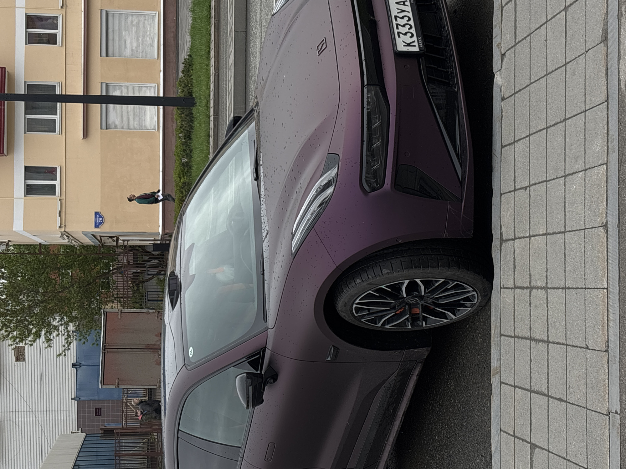 The image shows a purple car parked on a city street, possibly in Perm, Russia, based on the license plate "K333YA". The car is parked near a sidewalk paved with gray rectangular stones. In the background, there is a multi-story yellow building and some vegetation. A person is walking along the sidewalk in the distance. The weather appears to be overcast, as indicated by the droplets on the car.