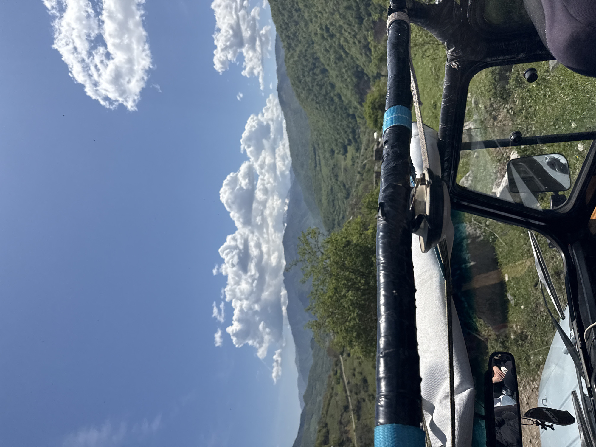 The image, likely taken from within a vehicle, shows a view of mountains and clouds against a blue sky. The location is inferred as Haghartsin, Armenia based on the presence of mountainous terrain and likely vegetation. The vehicle's frame, including a padded bar wrapped with black tape and blue accents, is visible. A white strap is connected to the bar. A side mirror and windshield wipers can be seen inside the car and a reflection of a person in the side mirror. The external landscape includes green slopes and cumulus clouds.