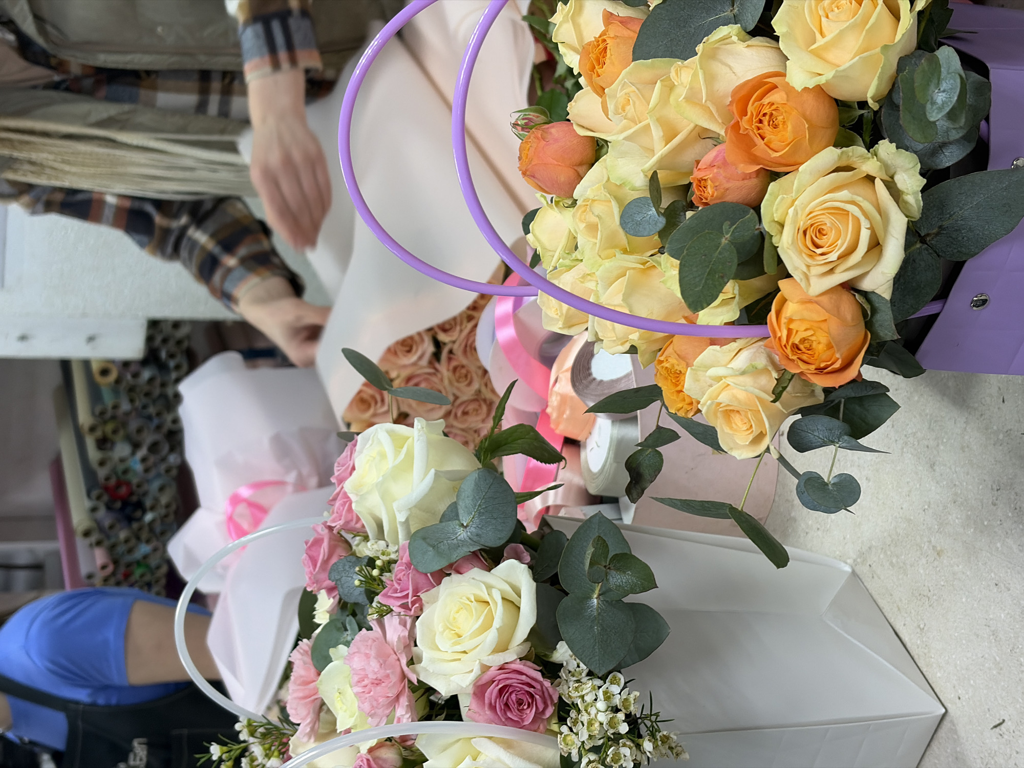 The image shows two bouquets of flowers, one featuring pale yellow and orange roses with eucalyptus leaves in a purple carrier with a matching handle, and the other with white and pink roses and eucalyptus leaves, alongside smaller white flowers, in a white carrier with a white handle. A person wearing a plaid shirt is arranging materials or working with flower packaging. Rolls of materials can be seen in the background. The location is likely a flower shop, potentially in Minsk, Belarus.