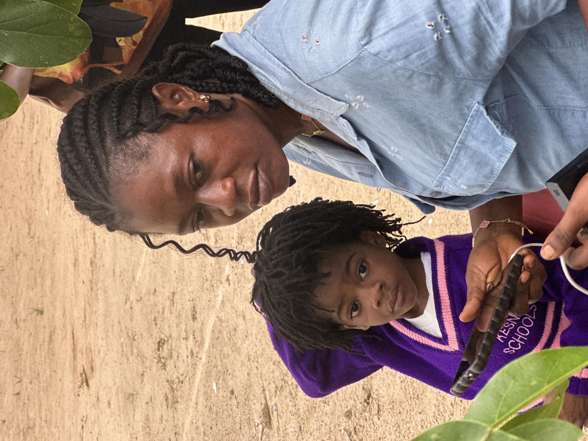 In Ganyingbo, Nigeria, a woman with braided hair and a light blue denim shirt sits next to a young child wearing a purple sweater. The woman is holding a dark-colored smartphone in her right hand, and a white charging cable is plugged into it. The child is looking directly at the camera. The background consists of a sandy, dirt-like ground with sparse vegetation and a blurred figure of another person partially visible behind the woman. The lighting suggests an outdoor setting, possibly during the daytime.