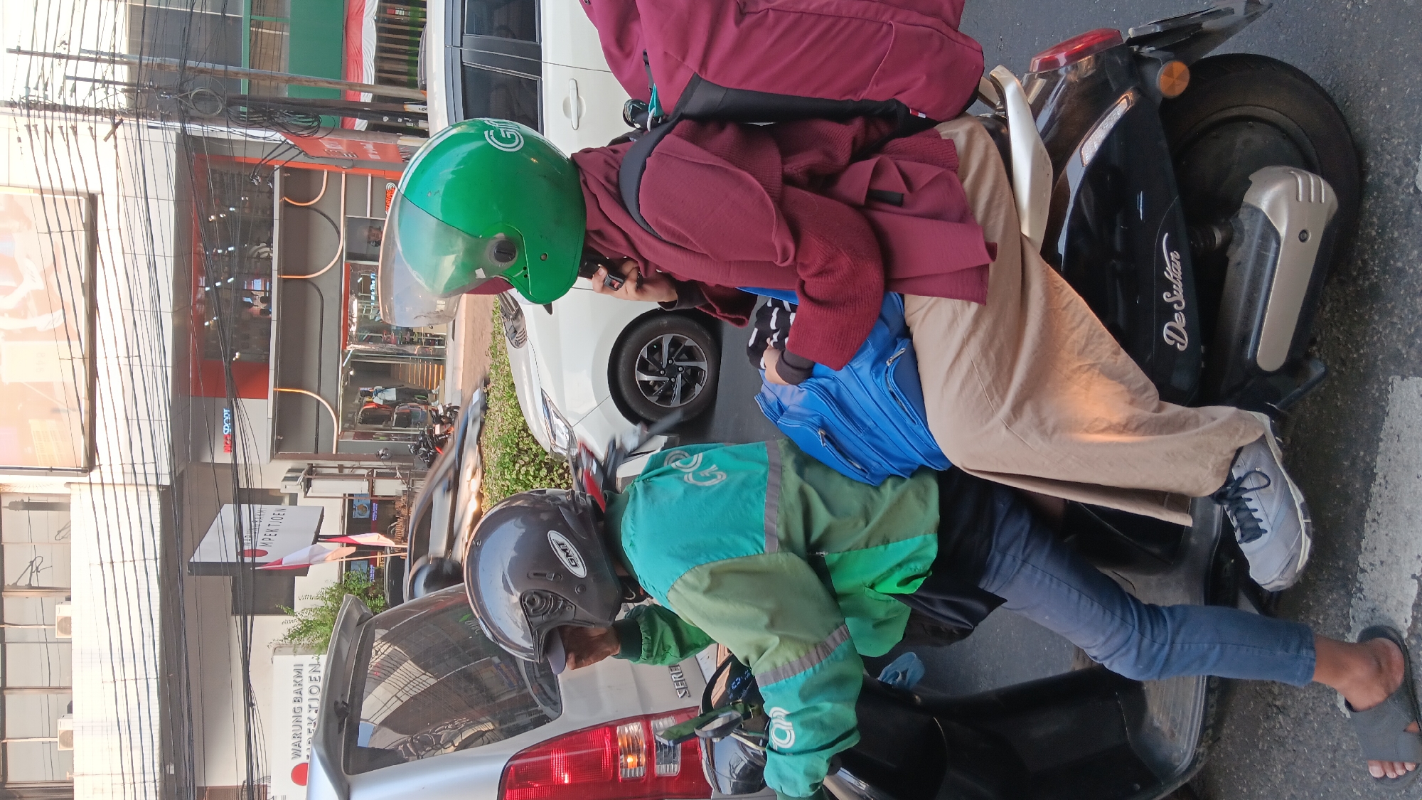Two individuals are riding on a black scooter in a street scene in South Jakarta, Indonesia. The person in front, wearing a green jacket with the Gojek logo and a grey helmet, is the driver. The passenger behind them wears a maroon jacket, a hijab, and a green helmet, and is speaking on a mobile phone. Both individuals have backpacks. The scooter is parked on a street with other vehicles and buildings visible in the background. The buildings appear to be commercial establishments, one of which has "SPORT" and "MPEKTJOEN" signage. The road is paved and marked with white lines. The lighting suggests it is daytime.