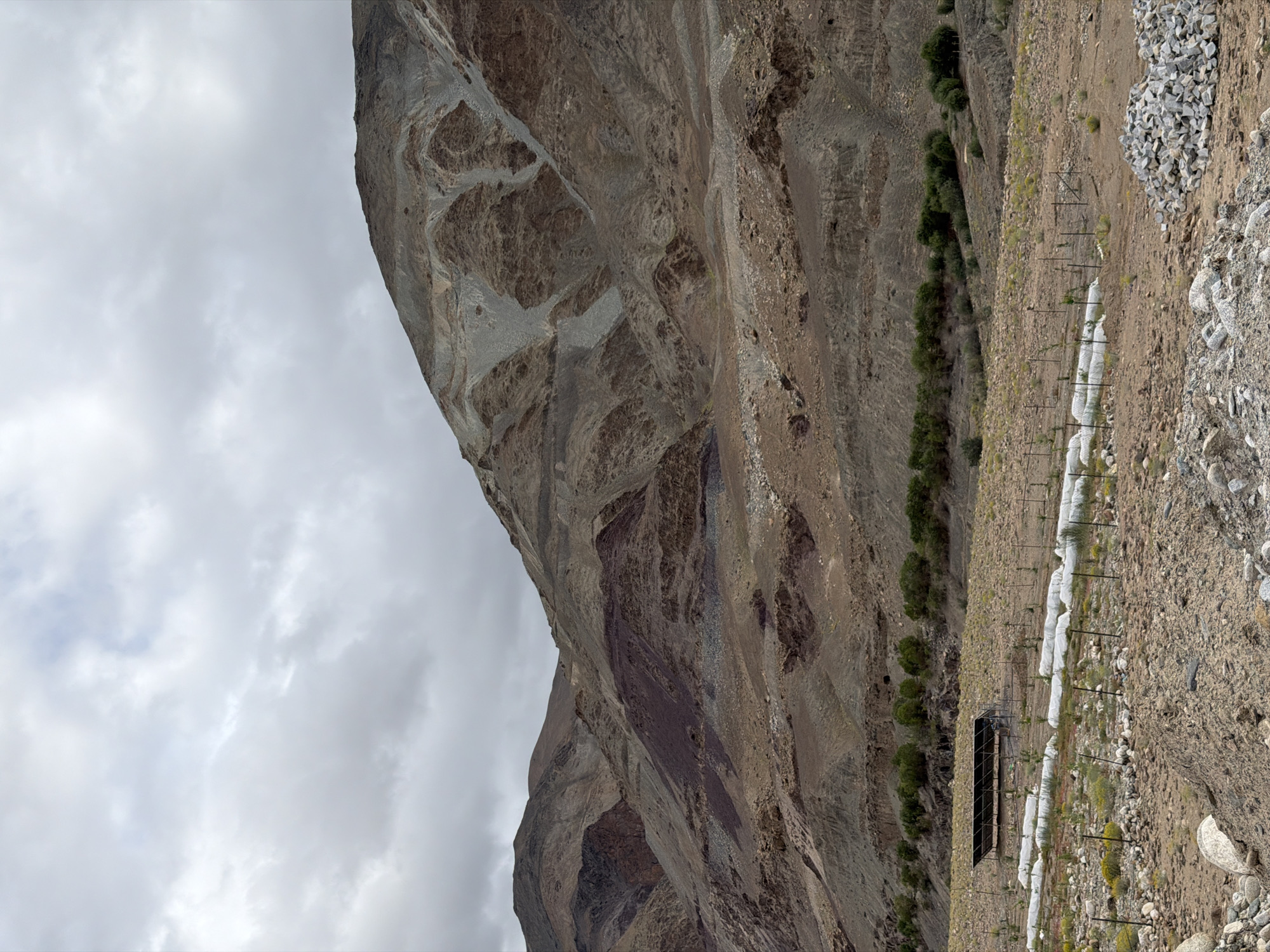 The image depicts a mountainous landscape in Kiari, India, under a cloudy sky. The foreground is a rocky, arid terrain with scattered vegetation. In the middle ground, there's a cultivated area featuring rows of plants covered in white protective material and a dark-colored structure, possibly a shade net or solar panel setup. A line of green trees borders this area. The background is dominated by large, barren mountains with visible layers of rock and soil, exhibiting shades of brown, gray, and reddish hues. The overall scene suggests a rural or remote location with agricultural activity in a harsh environment.