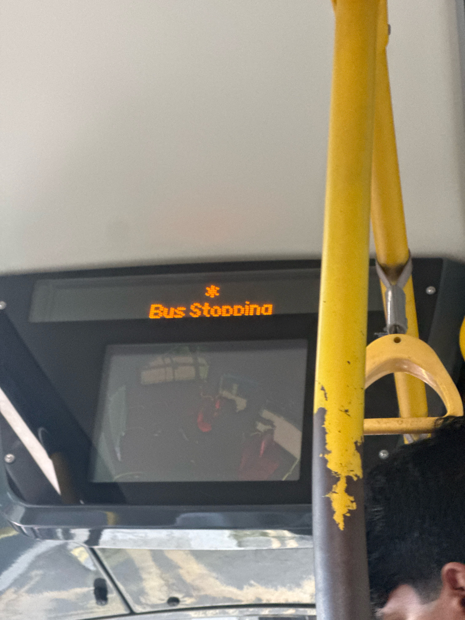 The image shows a digital display on a bus that reads "Bus Stopping" in orange LED text, with a snowflake-like icon to the left of the text. The display is mounted above a reflective screen, possibly showing passengers inside the bus. Yellow grab handles and poles are visible in the foreground, indicating the interior of a public transport vehicle. The overall scene suggests a bus in operation within the city of Greater London, United Kingdom.