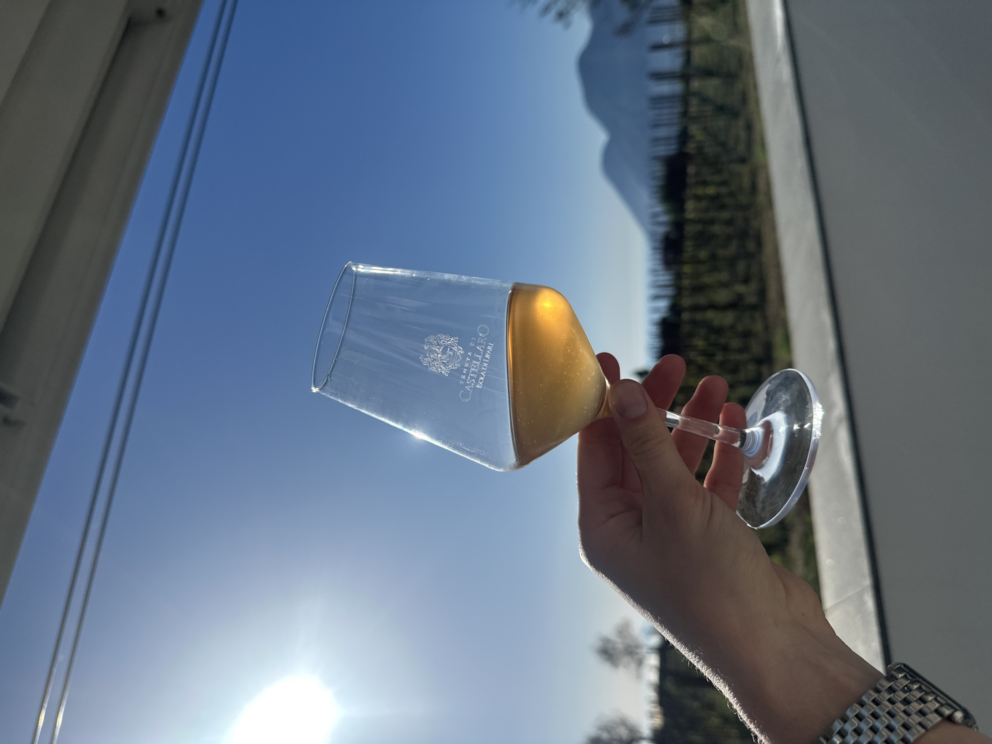 A close-up, angled shot captures a person's hand holding a clear champagne flute filled with a golden, sparkling beverage, likely champagne or sparkling wine. The glass bears the etched text "TENUTA DI CASTELLARO" and "ISOLA DI LIPARI," visible in reverse due to the glass's transparency. A metallic, checkered watch or bracelet adorns the wrist of the hand.
The setting is outdoors, beneath a vast, clear blue sky. The bright sun is prominently positioned in the lower left, creating a significant lens flare across the image. In the background, dark silhouettes of mountains or hills are visible against the sky, suggesting a rugged landscape. Below these natural formations, rows of what appear to be a vineyard or cultivated land are faintly discernible. Portions of a light-colored building or structure frame the scene on the left and right edges.
The image conveys a sense of enjoyment or celebration on a bright, sunny day, with the location cues on the glass specifically placing the event in Lipari, Italy. The activity is one of savoring a drink amidst a scenic backdrop, likely appreciating the natural beauty and the beverage.