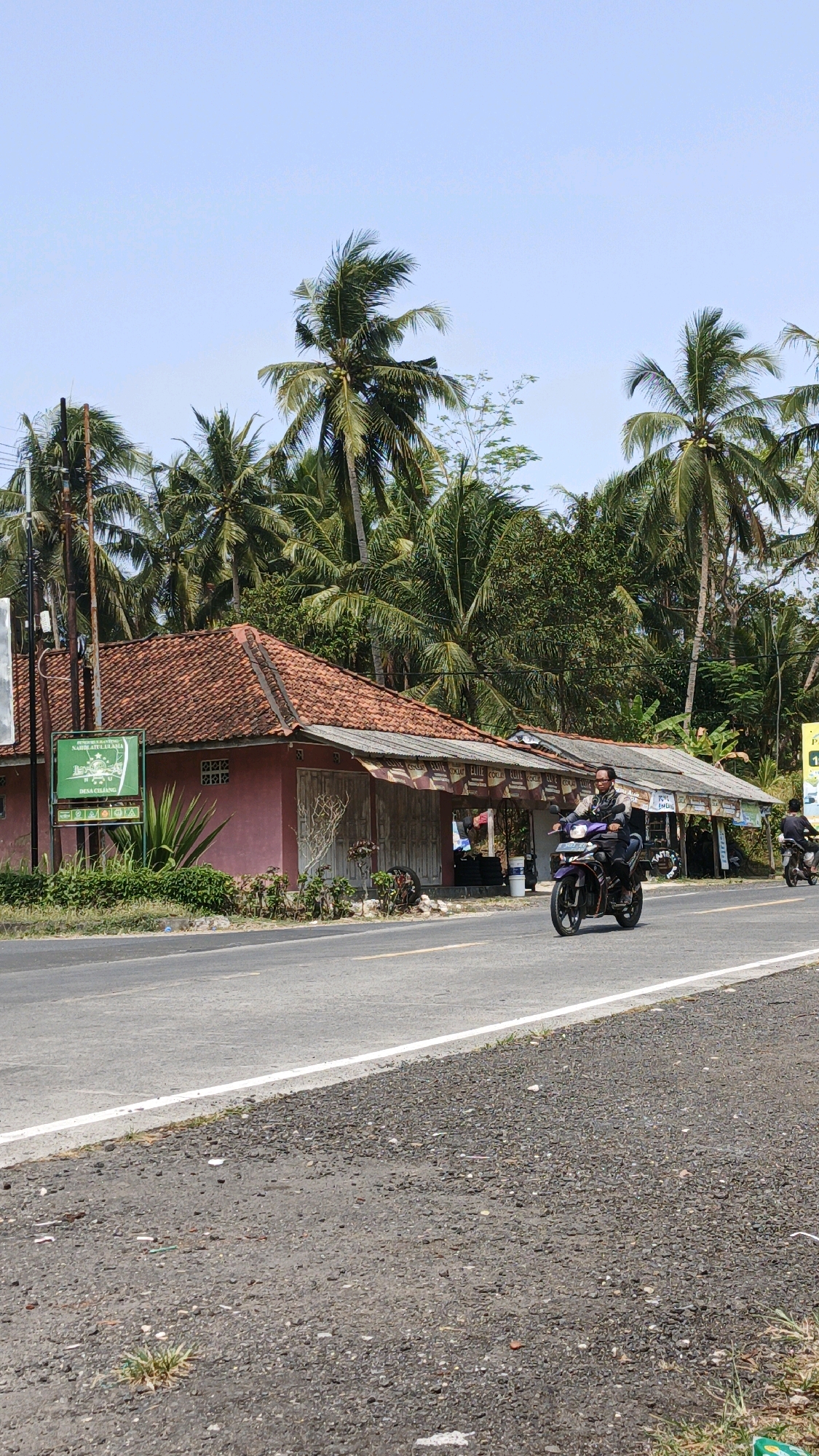 The image depicts a street scene in Cibenda, Indonesia, characterized by a bright, sunny day under a clear blue sky. A paved road runs horizontally across the foreground, with a white line marking its edge on the left. To the right of the road, the ground is covered in dark gravel and scattered debris. On the left side of the road, there is a building with a red tiled roof. This building is pink and appears to be a shop or small establishment, with white shutters and a sign on the left side. Lush green foliage, including palm trees, forms a dense backdrop behind the building.
In the middle ground, a man is riding a dark-colored motorcycle, moving from left to right along the road. He is wearing a patterned jacket and a helmet. Another motorcyclist, further down the road and less visible, is also moving in the same direction. The shops lining the road appear to have awnings and various signs. The overall atmosphere suggests a warm, tropical environment.
