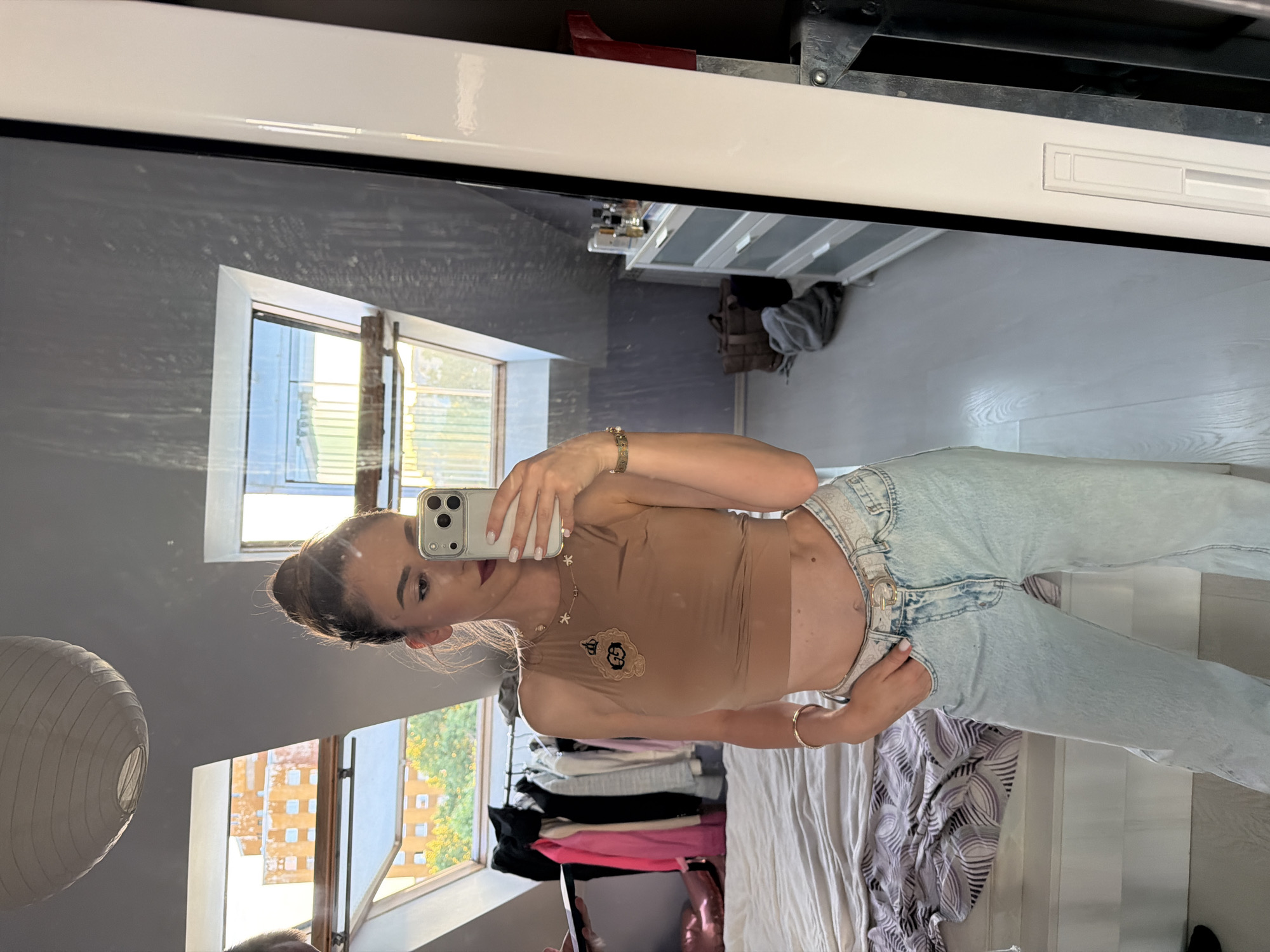 A young woman with her hair pulled up in a bun, wearing a tan crop top and light blue jeans, stands in a room and takes a mirror selfie with her phone. She is wearing a gold necklace with star charms and a gold bracelet on her left wrist. The room appears to be a bedroom with a bed, a clothing rack with several items of clothing, and a dresser. There are also several skylights in the ceiling, allowing natural light to enter the room. The image shows a partial view of a brick building through one of the skylights. Based on the architectural style of the building visible through the skylight, it's likely this scene is located in Sofia, Bulgaria.