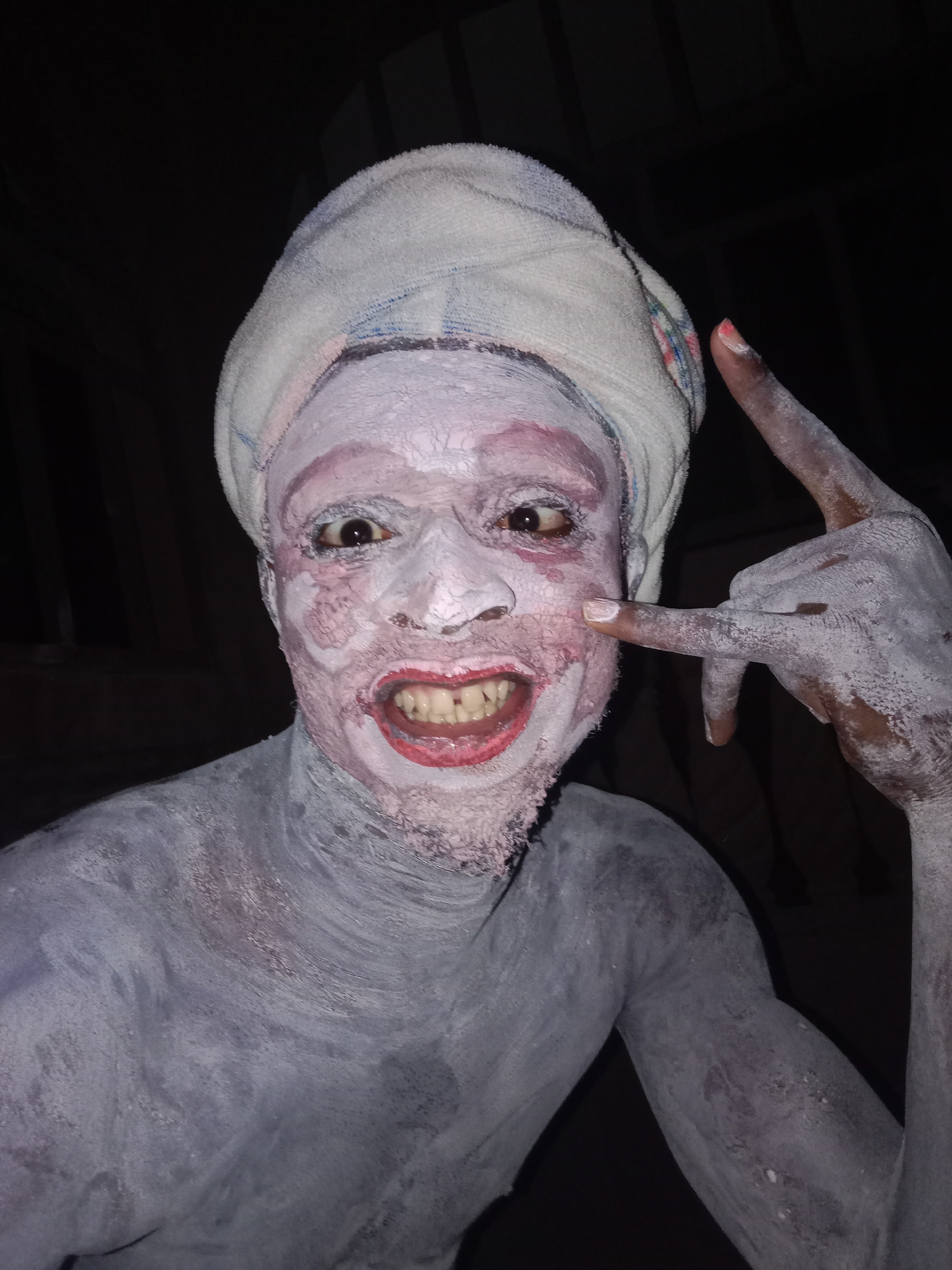A medium shot depicts a person heavily covered in white and light gray powder, possibly clay or a similar substance. They are wearing a white towel wrapped around their head. The person is making a gesture with their right hand, index and pinky fingers extended, while the other fingers are curled inward. Their face is painted with pink around the eyes and lips, and their teeth are visible in a broad, somewhat distorted smile. The background is dark and indistinct, with some blurred shapes suggesting an interior space. The location is Unknown, Unknown.