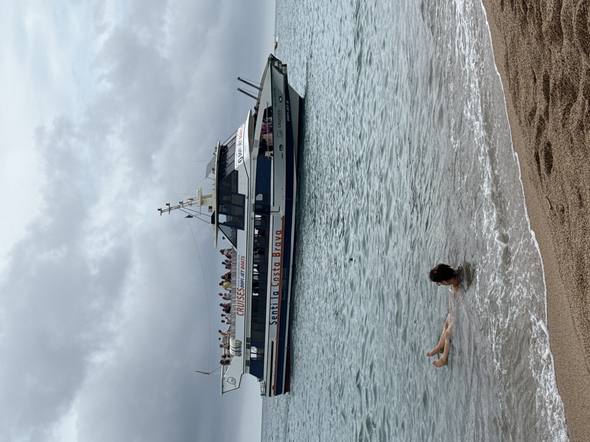The image depicts a cloudy day at the beach in Blanes, Spain. In the foreground, a person is partially submerged in the shallow, wavy water, with their feet and lower legs visible. The water laps at a sandy beach, which occupies the bottom right of the frame. In the mid-ground and background, a white and blue boat is sailing on the sea. The boat, identified by text as "Senti la Costa Brava" and "DOFI JET BOATS," carries numerous passengers on its upper deck. The sky is overcast with dramatic clouds.