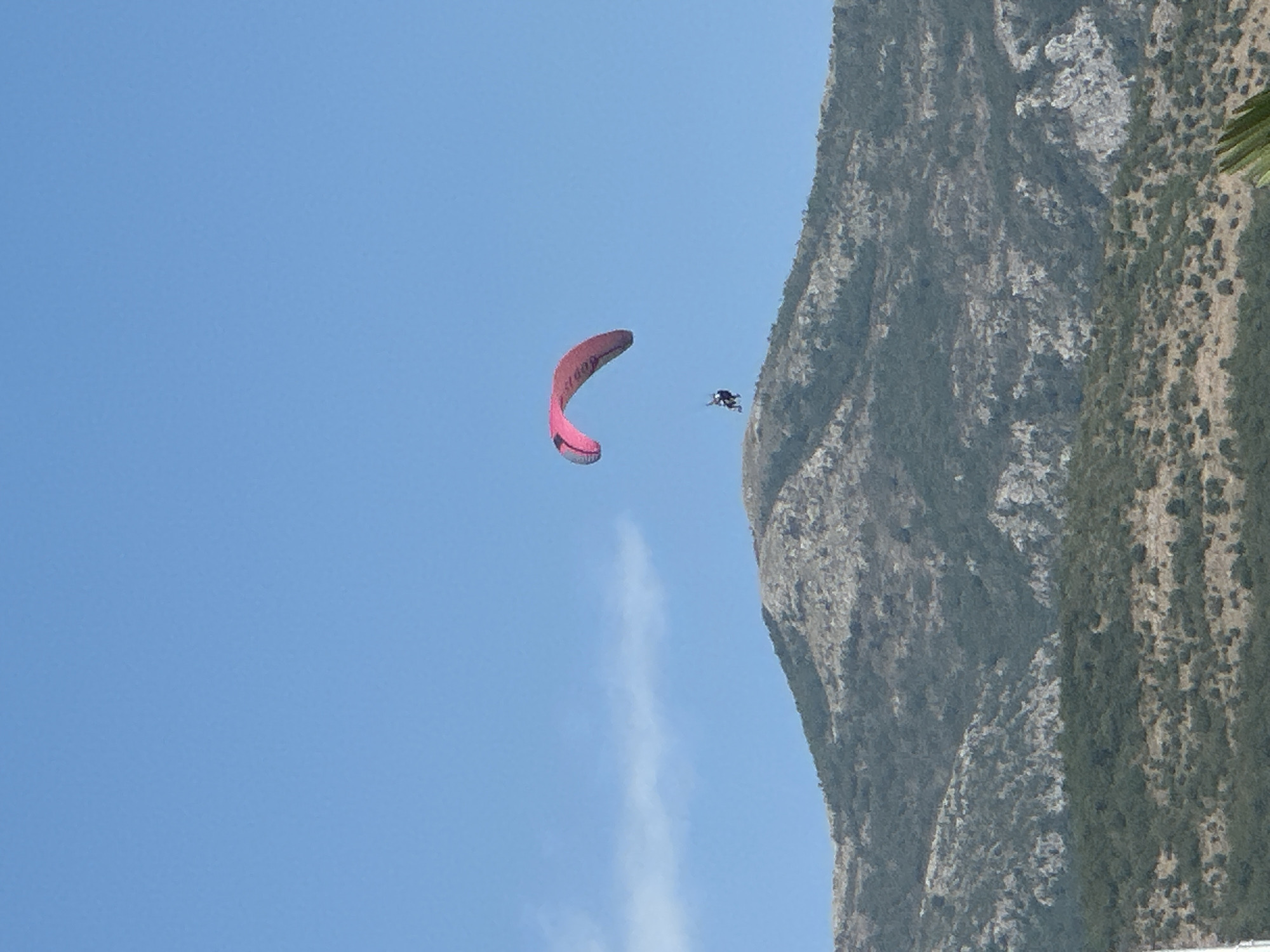 A person is paragliding over a mountainous terrain in Bečići, Montenegro. The paraglider, with a pink and grey wing, is suspended in the bright blue sky above a rugged, sparsely vegetated mountain. The mountain slope features a mix of green shrubbery and rocky outcrops. A faint wisp of cloud is visible in the sky.