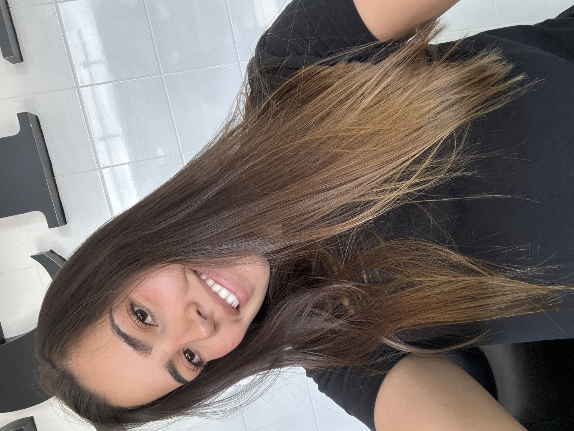 A cheerful young woman is captured in a portrait-style selfie, smiling broadly and looking directly at the camera. She has long, dark brown hair that transitions to a lighter, caramel or light brown shade at the ends, consistent with an ombre or balayage style. Her hair flows over her shoulders and chest. She has dark, full eyebrows and dark eyes, and her wide smile reveals white teeth. The woman is wearing a simple black top or t-shirt. The background features a white tiled wall with distinct grout lines, suggesting an indoor environment such as a bathroom, kitchen, or salon. Partially visible dark, geometric objects, possibly furniture or architectural elements, are present on the left side of the frame. The lighting is bright and even, indicating an indoor setting in San Bernardino Tlaxcalancingo, Mexico. There is no visible text.