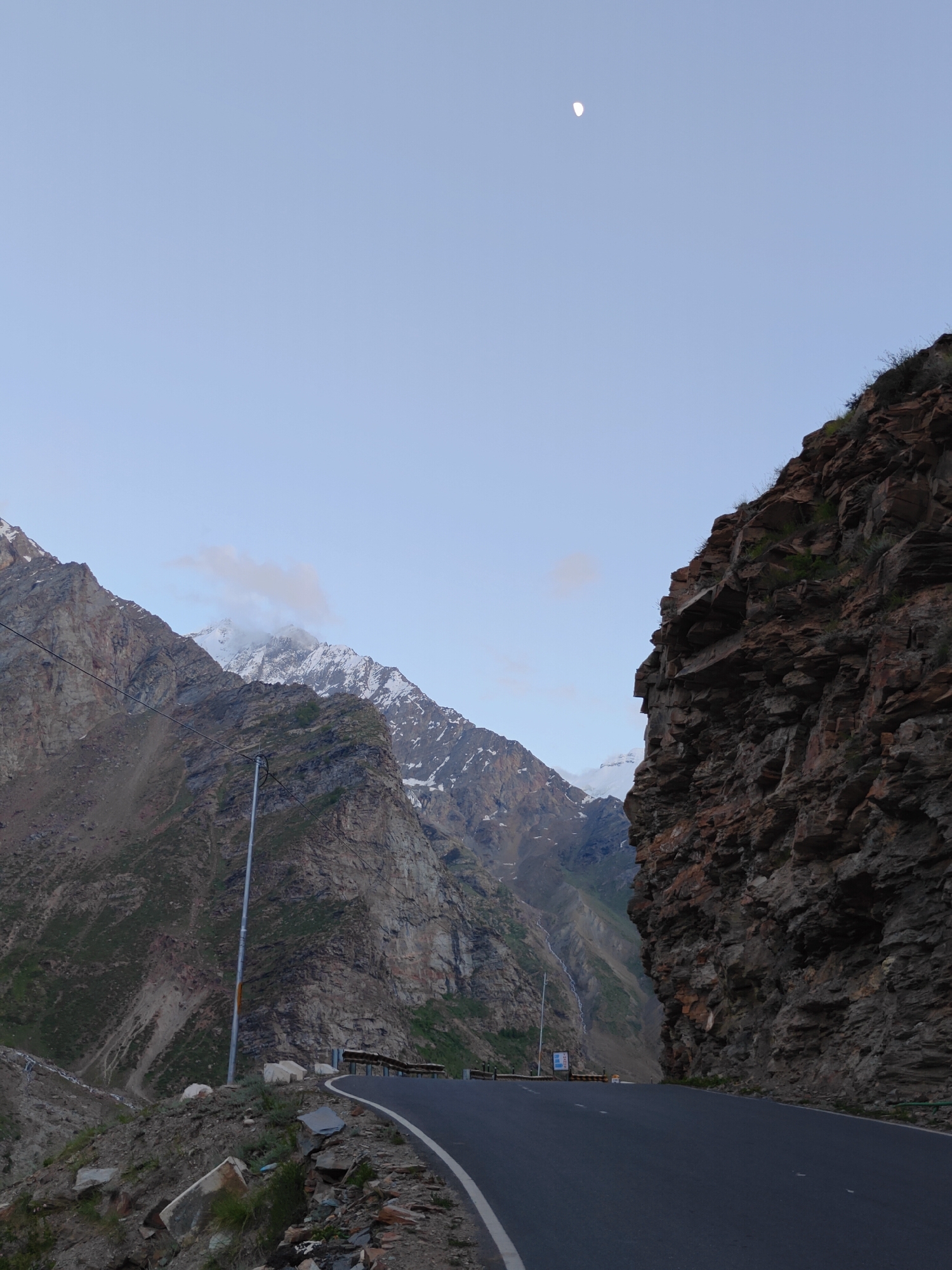 The image depicts a scenic mountain road in Gemur, India, at dusk. The sky is a pale blue with a sliver of the moon visible. To the right, a sheer rock face dominates the foreground, its rough texture and reddish-brown hues contrasting with the muted tones of the landscape. A winding asphalt road curves through the scene, marked by a white line. On the left, imposing mountains rise, their slopes covered in a mix of rock and sparse vegetation. Snow-capped peaks are visible in the distance, partially obscured by clouds. A solitary streetlamp stands on the side of the road, casting a faint glow. The overall impression is one of a remote and rugged mountain pass, bathed in the soft light of twilight.